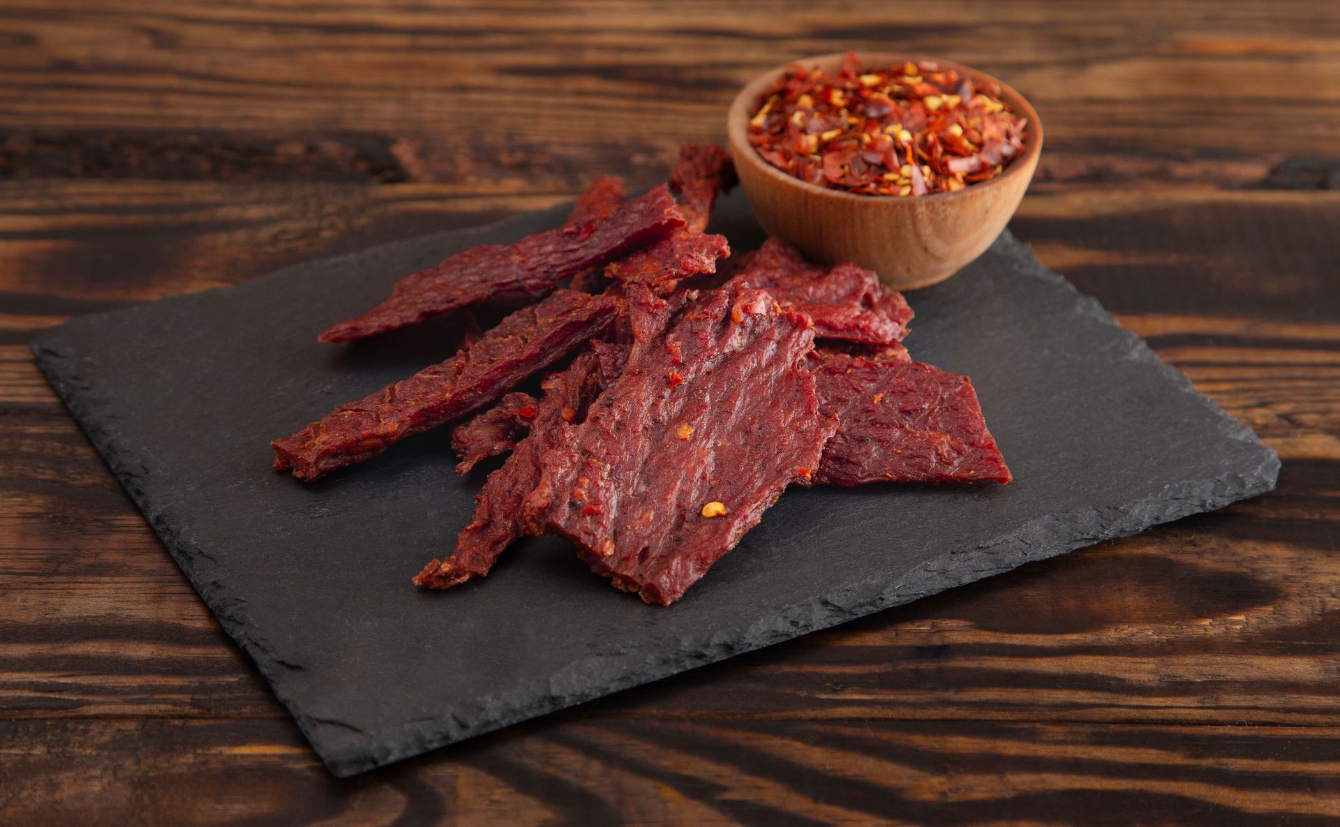 Beef jerky and chili flakes on a dark slate, wooden background.