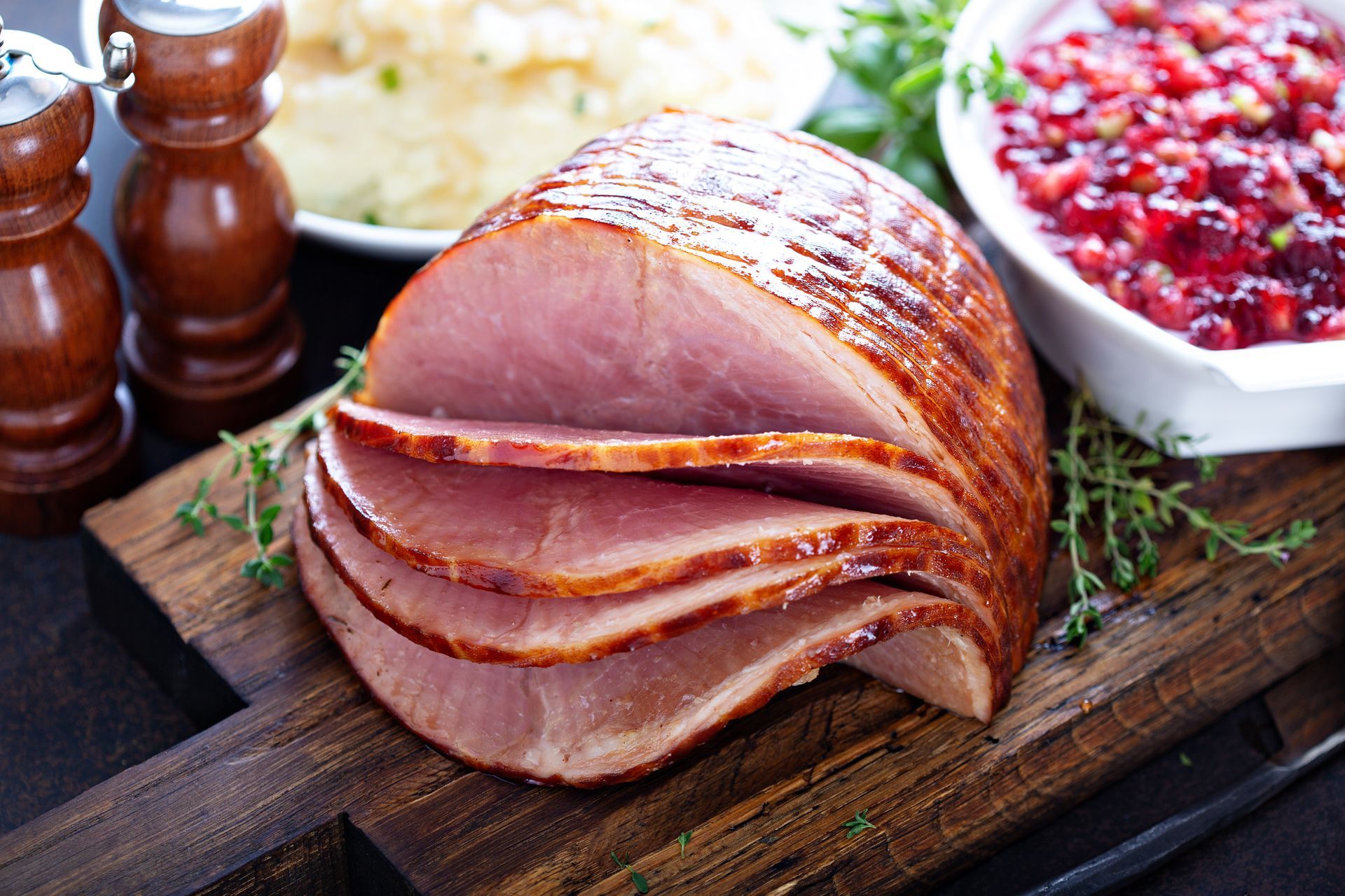 Sliced glazed ham on wooden cutting board, with cranberry sauce and mashed potatoes in background. Sliced glazed ham on wooden cutting board, with cranberry sauce and mashed potatoes in background.
