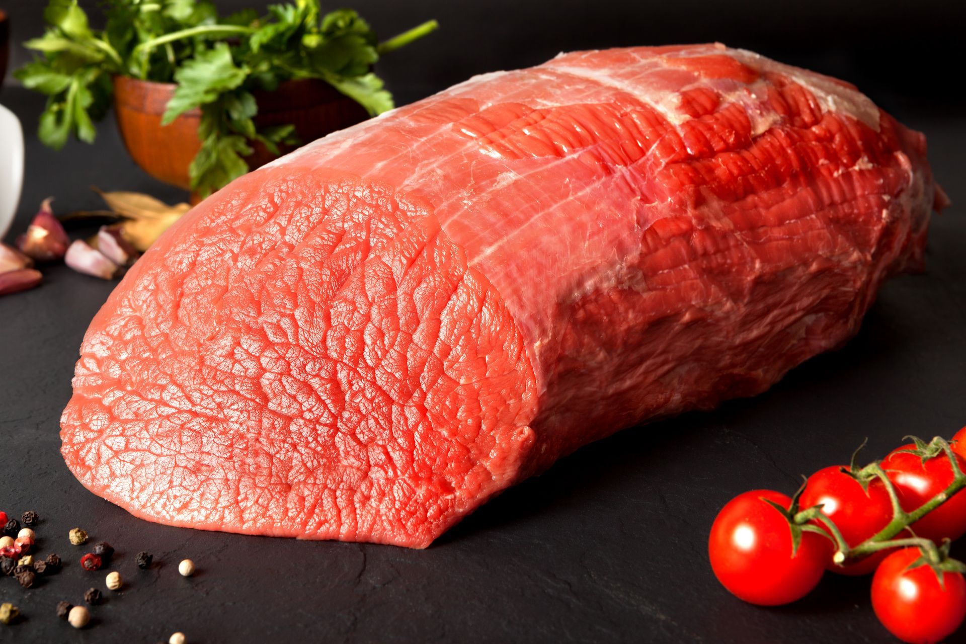 Raw beef roast, red with visible grain, surrounded by tomatoes, herbs, and spices on a black surface. Raw beef roast, red with visible grain, surrounded by tomatoes, herbs, and spices on a black surface.