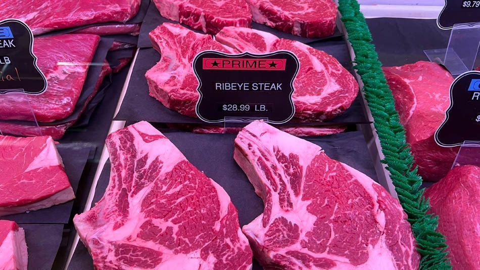 Display case of raw ribeye steaks with marbled fat, labeled as $26.99/lb. Display case of raw ribeye steaks with marbled fat, labeled as $26.99/lb.
