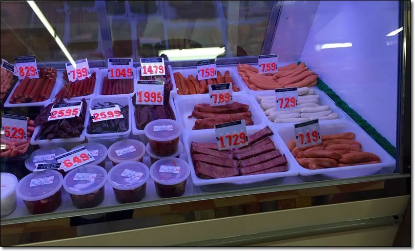 Meat display case in a store, filled with various sausages and prepared meats, priced with white labels.