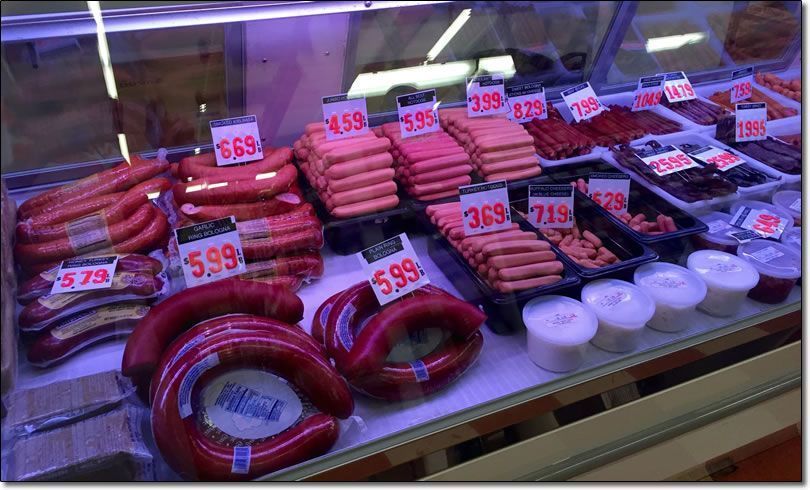 Butcher shop display with various sausages and prepared foods, priced.