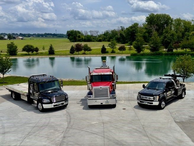 Three tow trucks are parked in a driveway next to a lake.