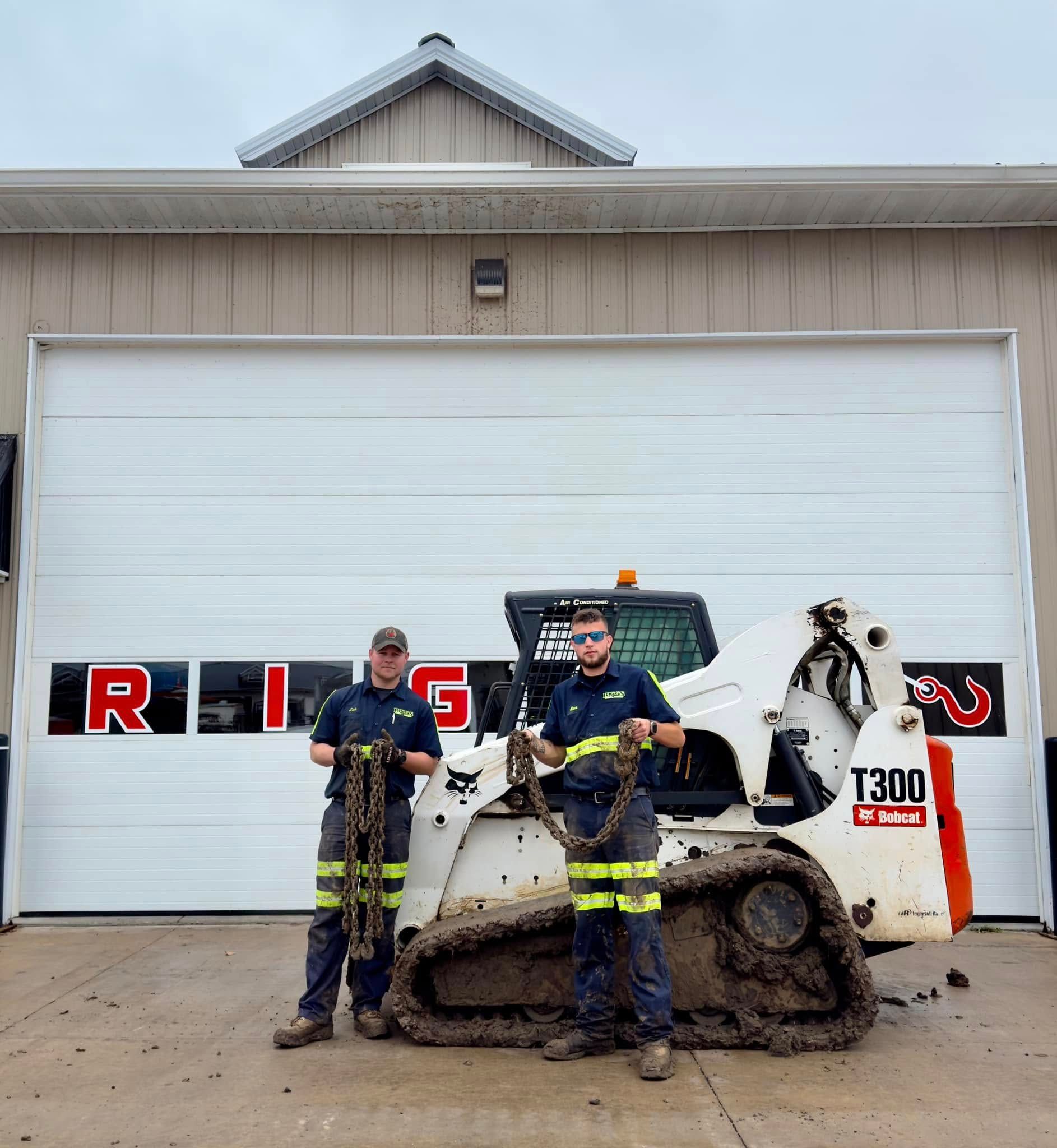 Two men are standing next to a bobcat in front of a garage door.