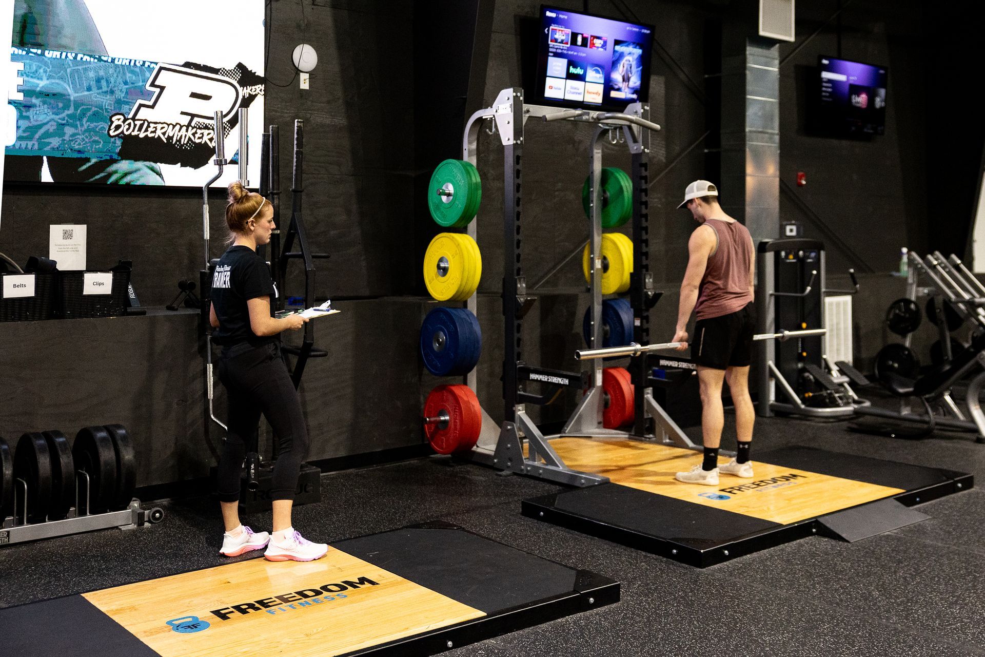 Member lifting a barbell while a personal trainer provides guidance in the Freedom Fitness weight room.