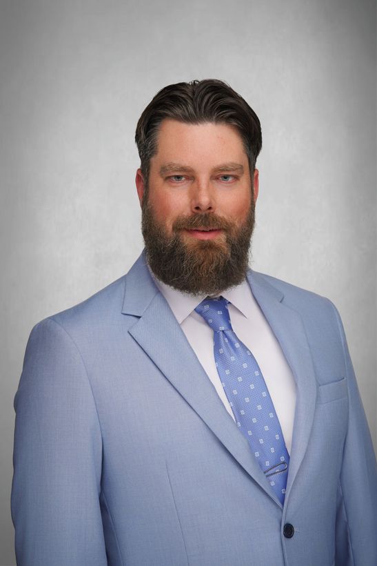 A man wearing a blue suit jacket and tie with a white dress shirt. The man has a welcoming smile. Grey background.