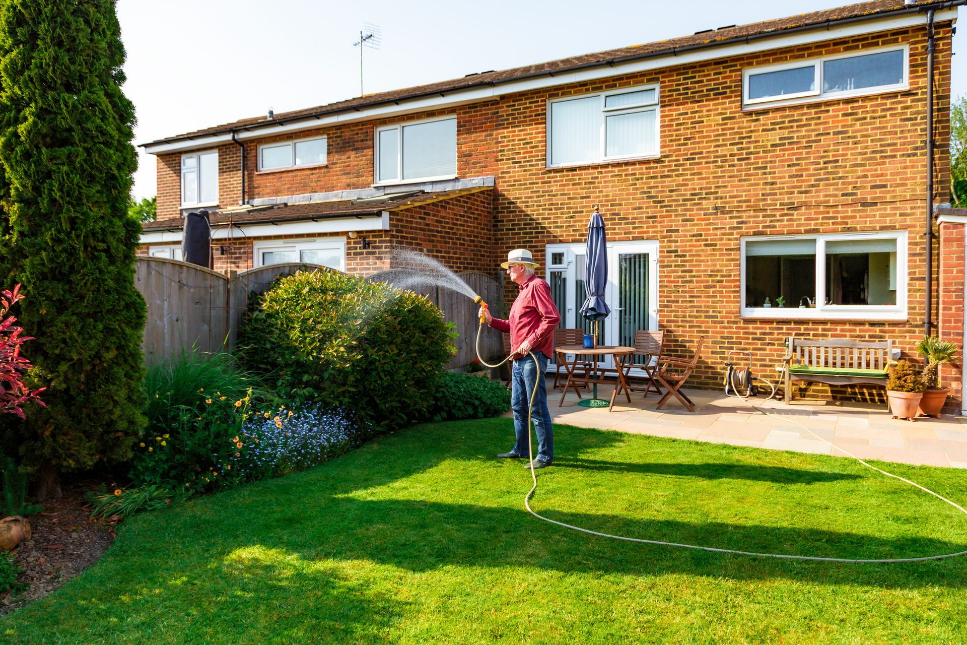 A person waters a garden in a sunny backyard with a brick house.