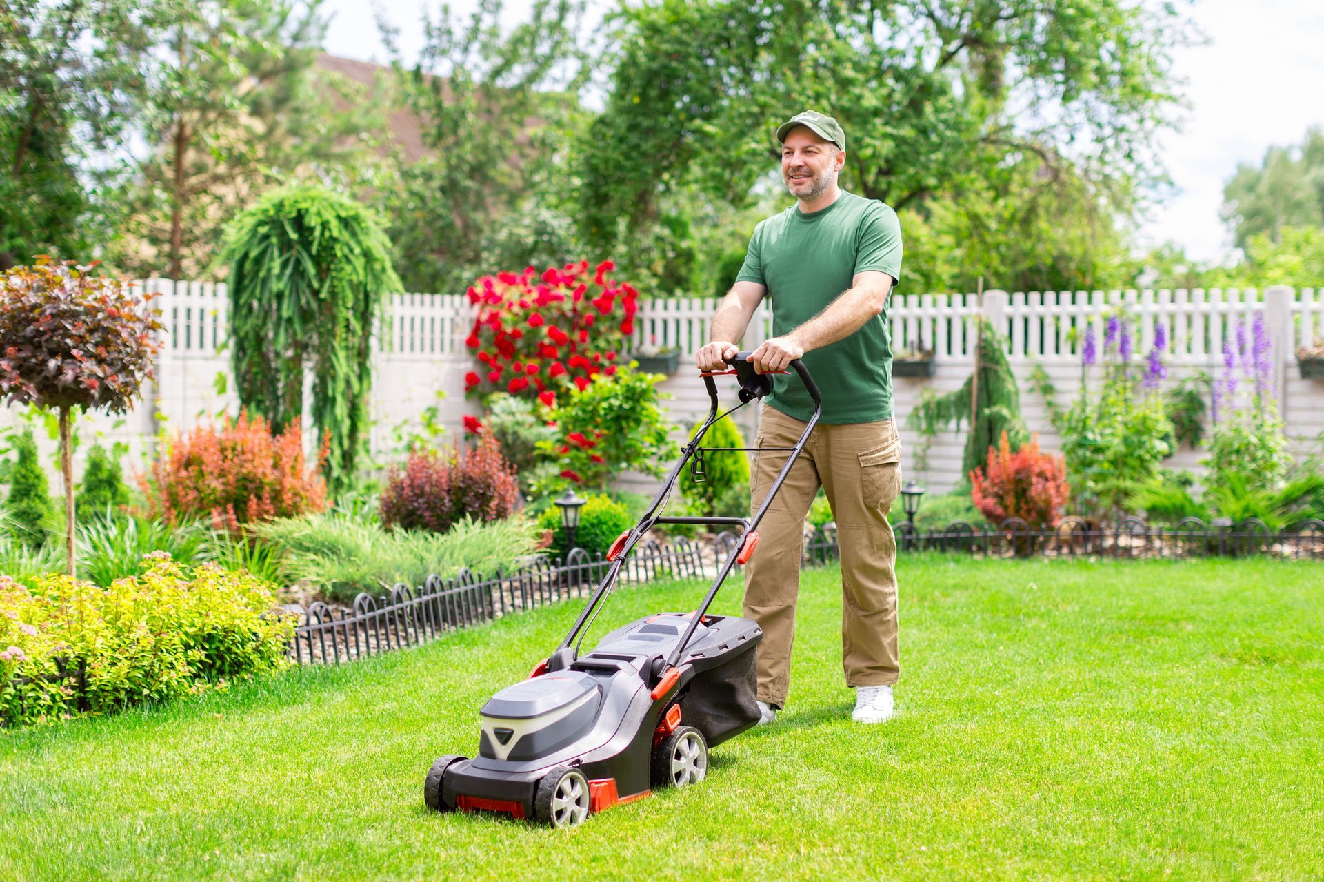 Man mowing the lawn in a well-kept garden with a white fence and colorful plants; sunny day.