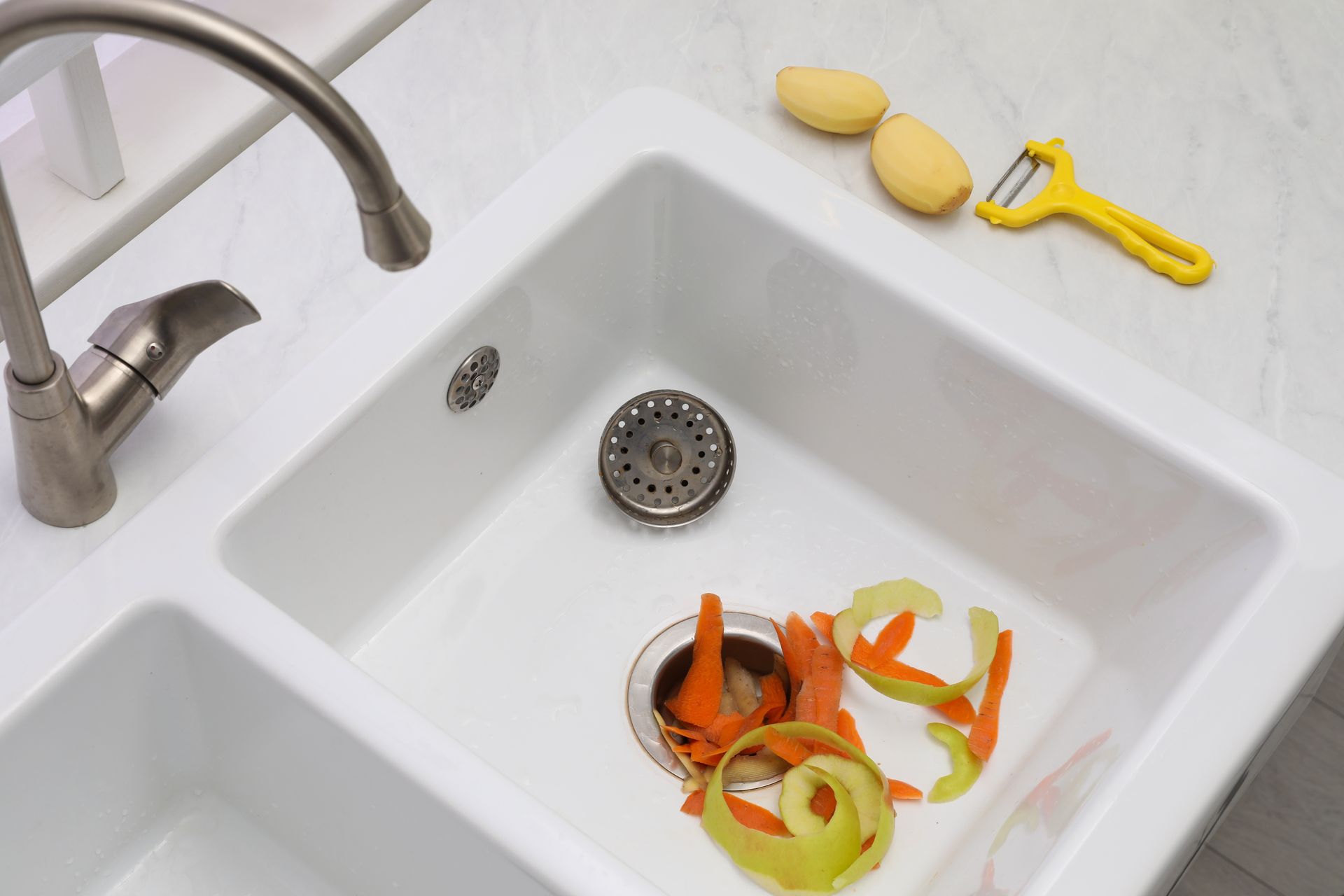 White kitchen sink with food scraps, potatoes, and a peeler.