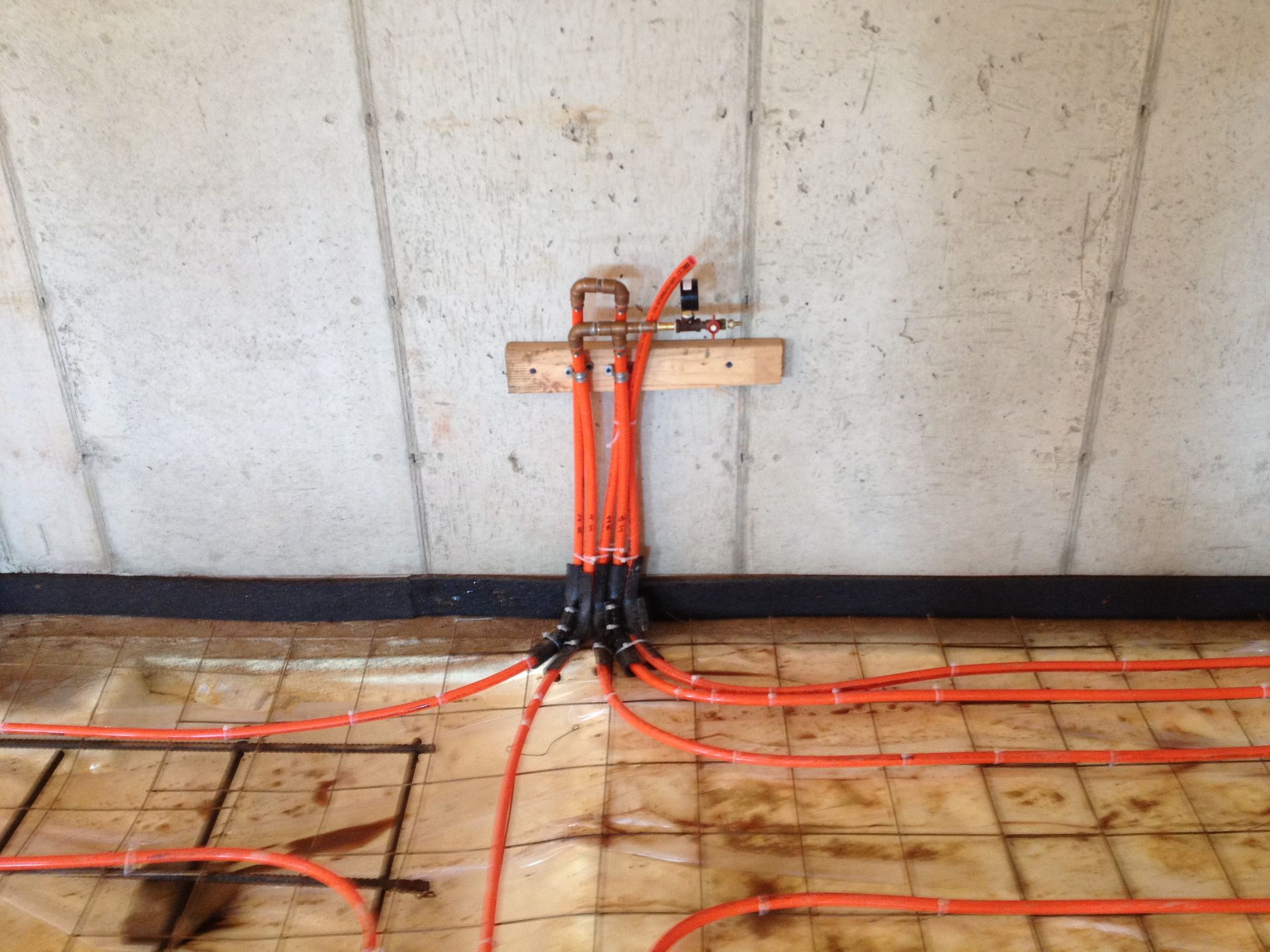 Orange radiant heating pipes attached to a concrete wall with wooden support and floor grid.