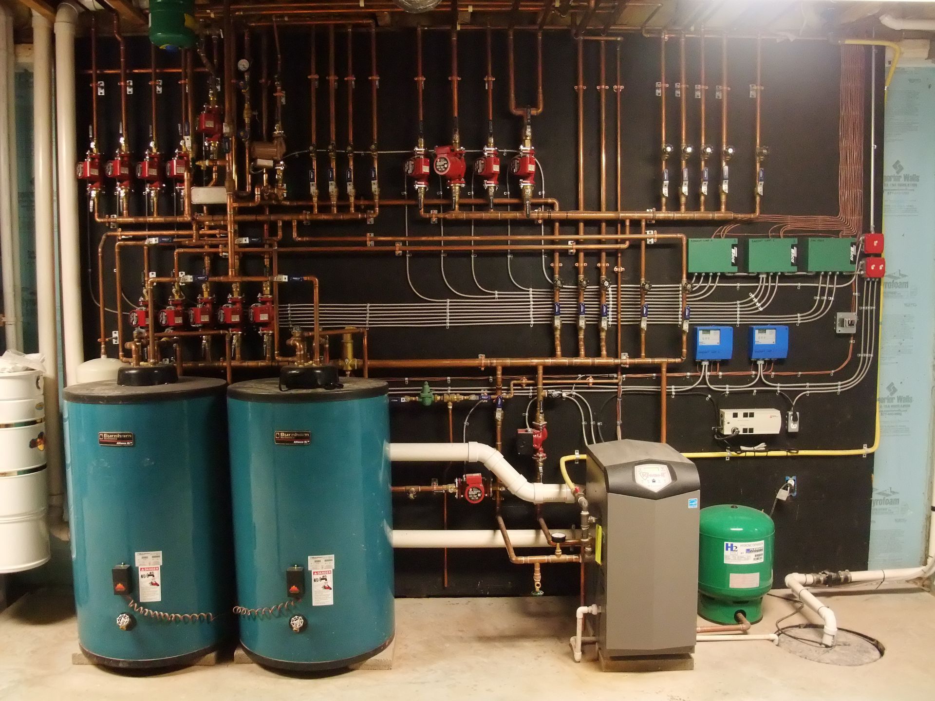 Two teal water heaters and complex copper piping on a black wall. A modern furnace is on the right.