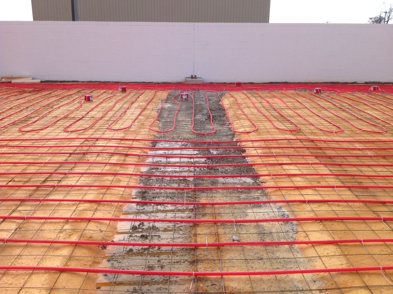 Construction site showing red radiant heating tubes laid on a surface, ready for concrete, near a white wall.