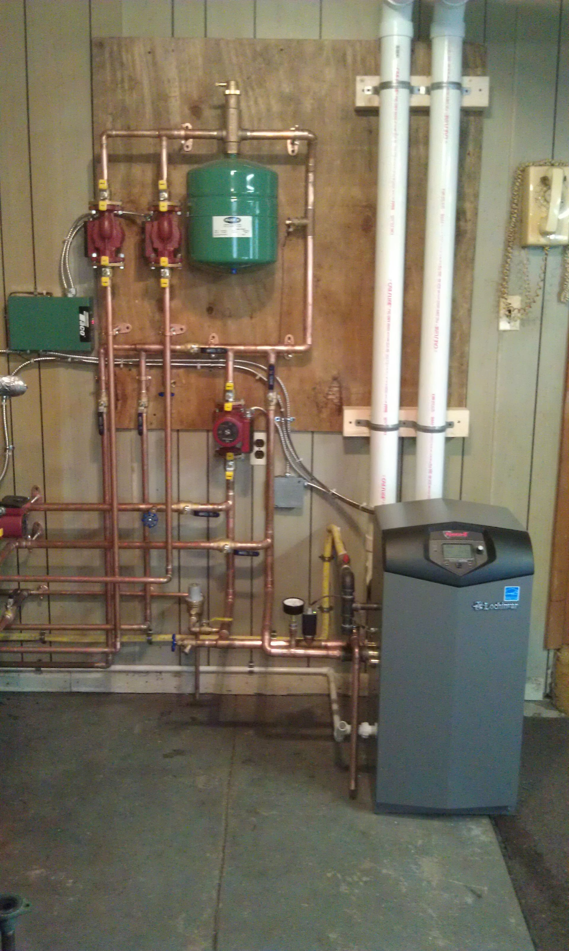 Heating system with copper pipes, green tank, pumps, and a gray boiler against a wooden wall.