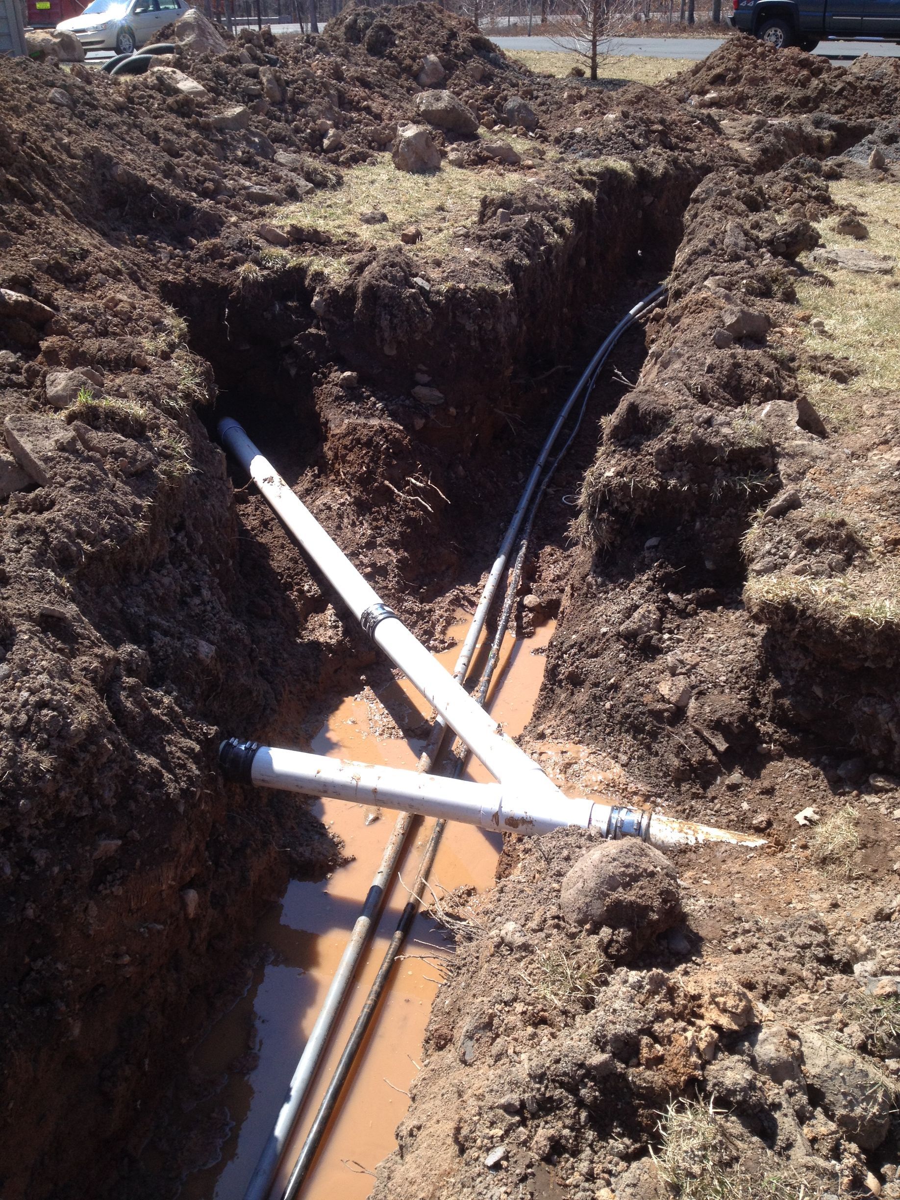 Trench with exposed white pipes, connecting and branching. Dirt walls and muddy water visible.