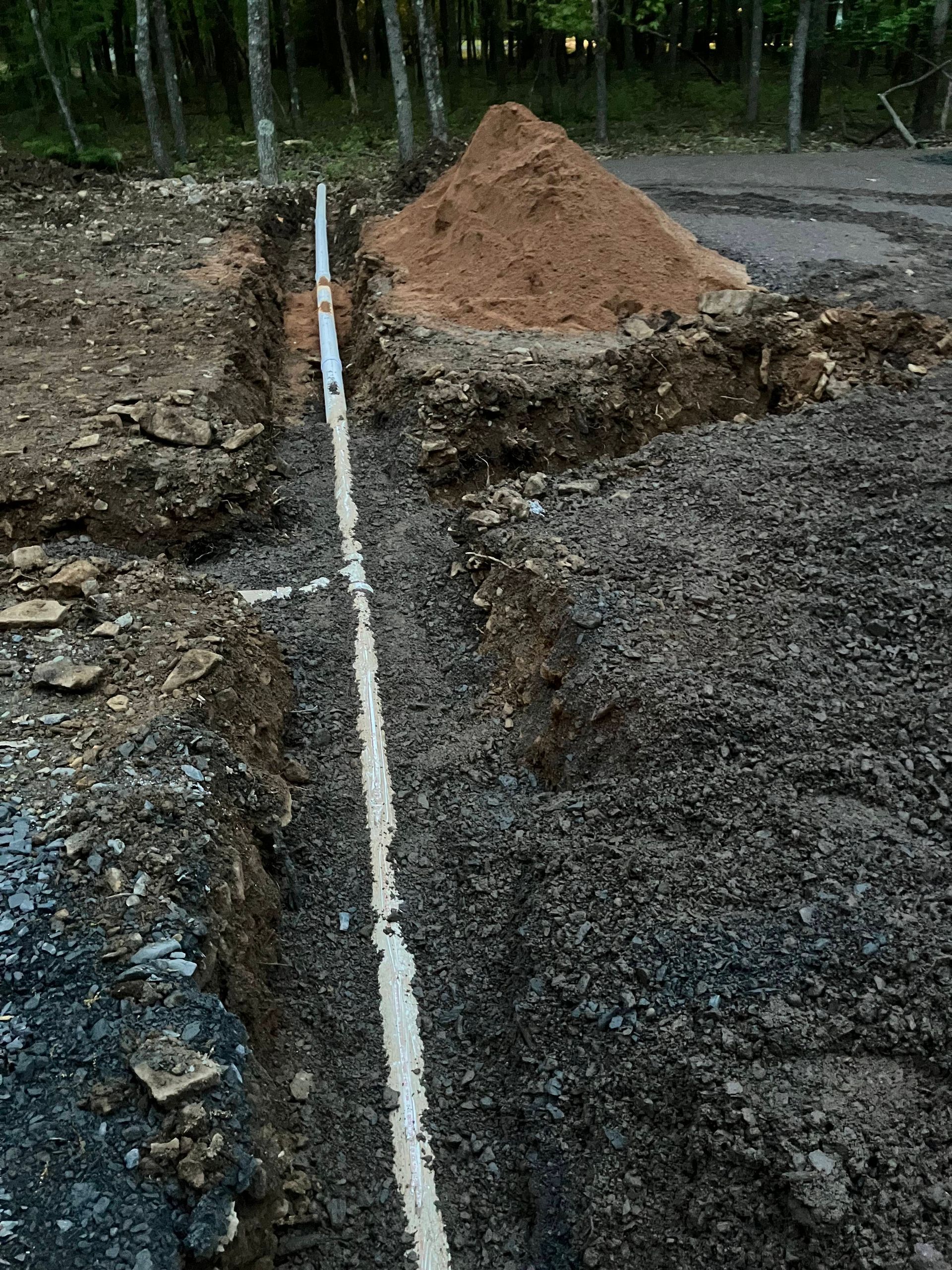 Trench dug for pipe; white pipe visible in the trench, with a pile of dirt and trees in the background.