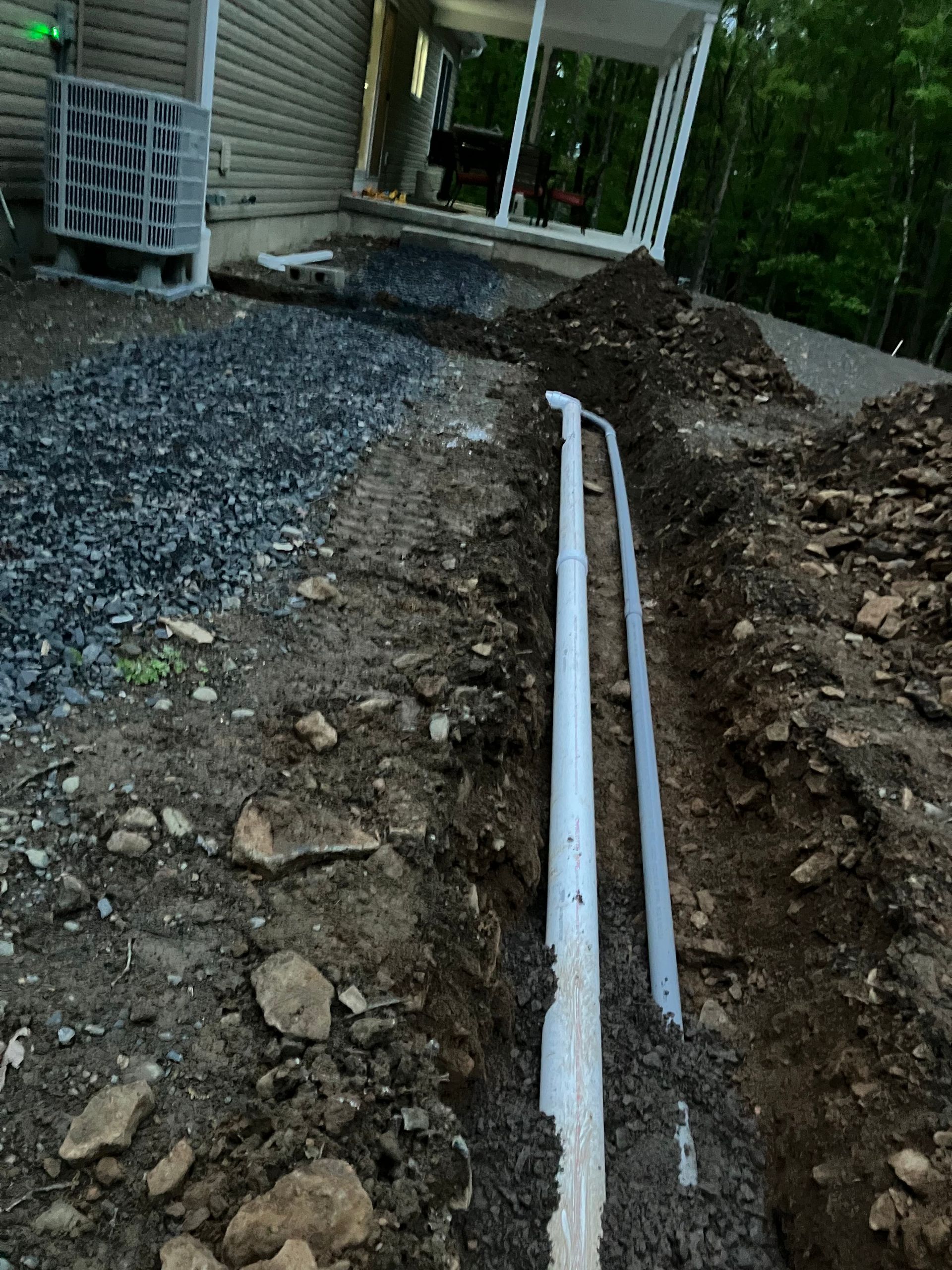 A trench in dirt with two white PVC pipes leading away from a house's foundation.