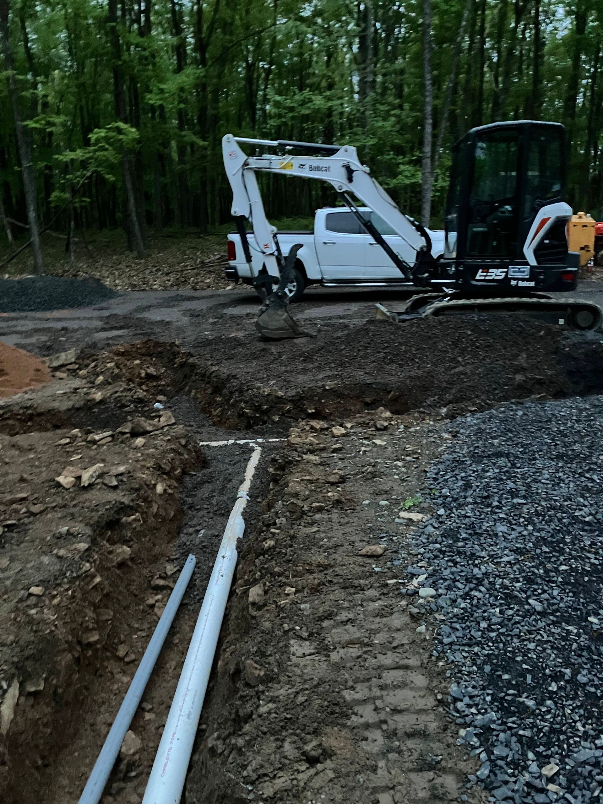 Trench with white PVC pipes and gray conduit; small excavator and white truck on construction site.