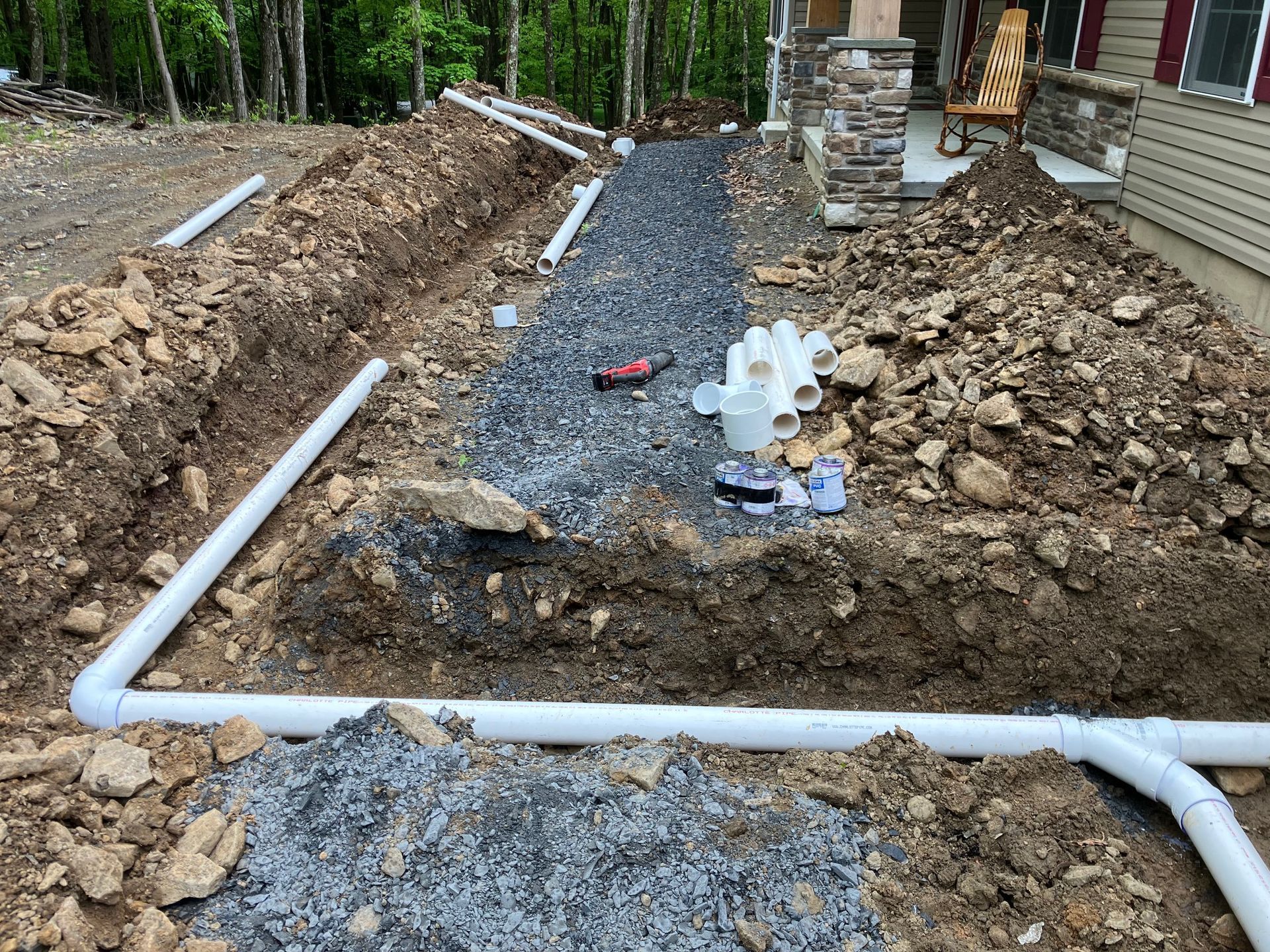 Construction site: Trench with gravel and white pipes leading to a house with stone columns.