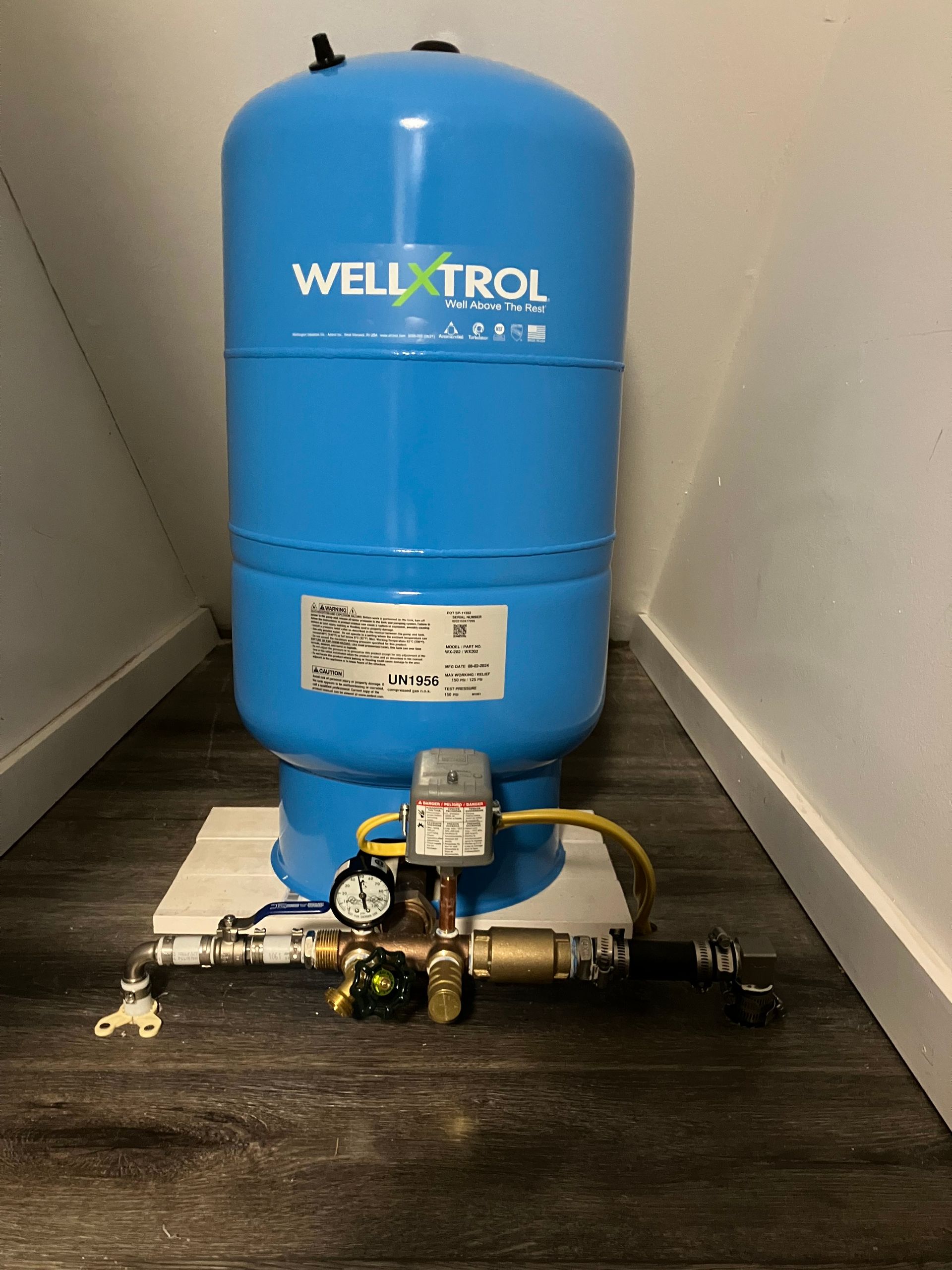 Blue WELL-X-TROL water pressure tank with associated plumbing on a wood floor in an enclosed space.