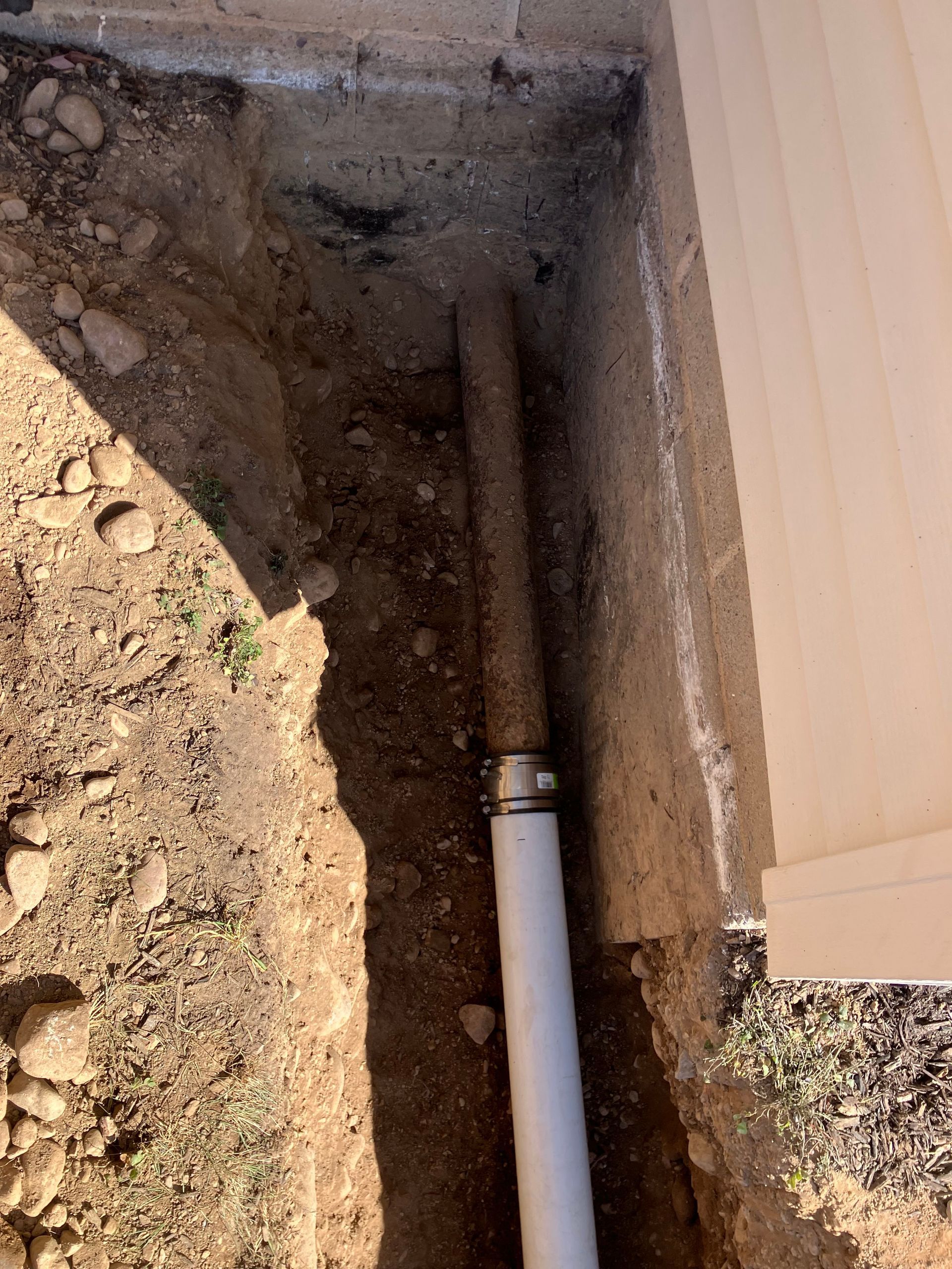 Trench with a white pipe and a metal fitting. Next to a beige wall, in the dirt.
