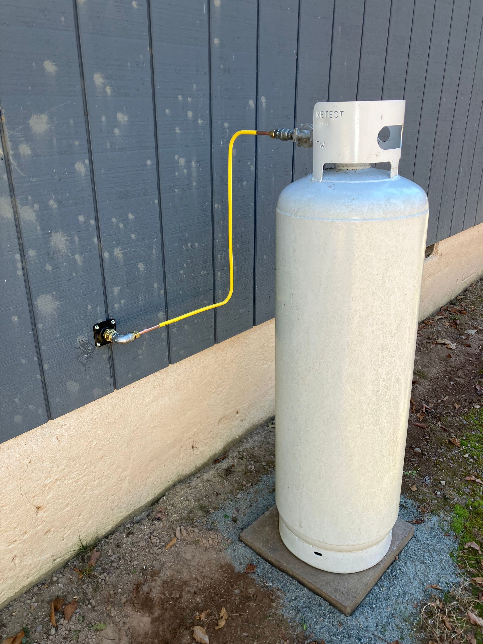 White propane tank with yellow gas line connected to a gray house wall.
