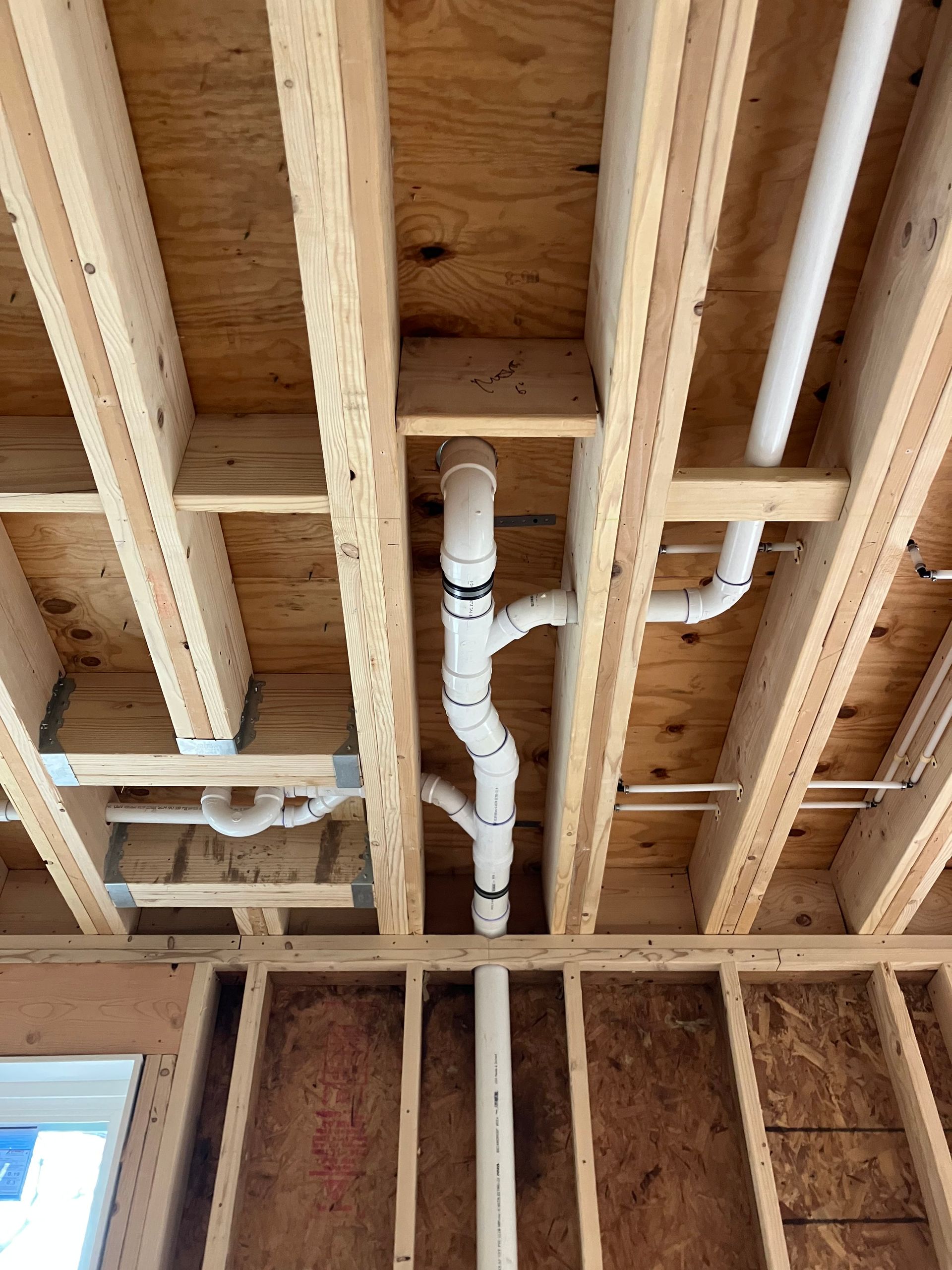 Piping installed in a wood frame ceiling and wall. White PVC pipes are connected for plumbing.