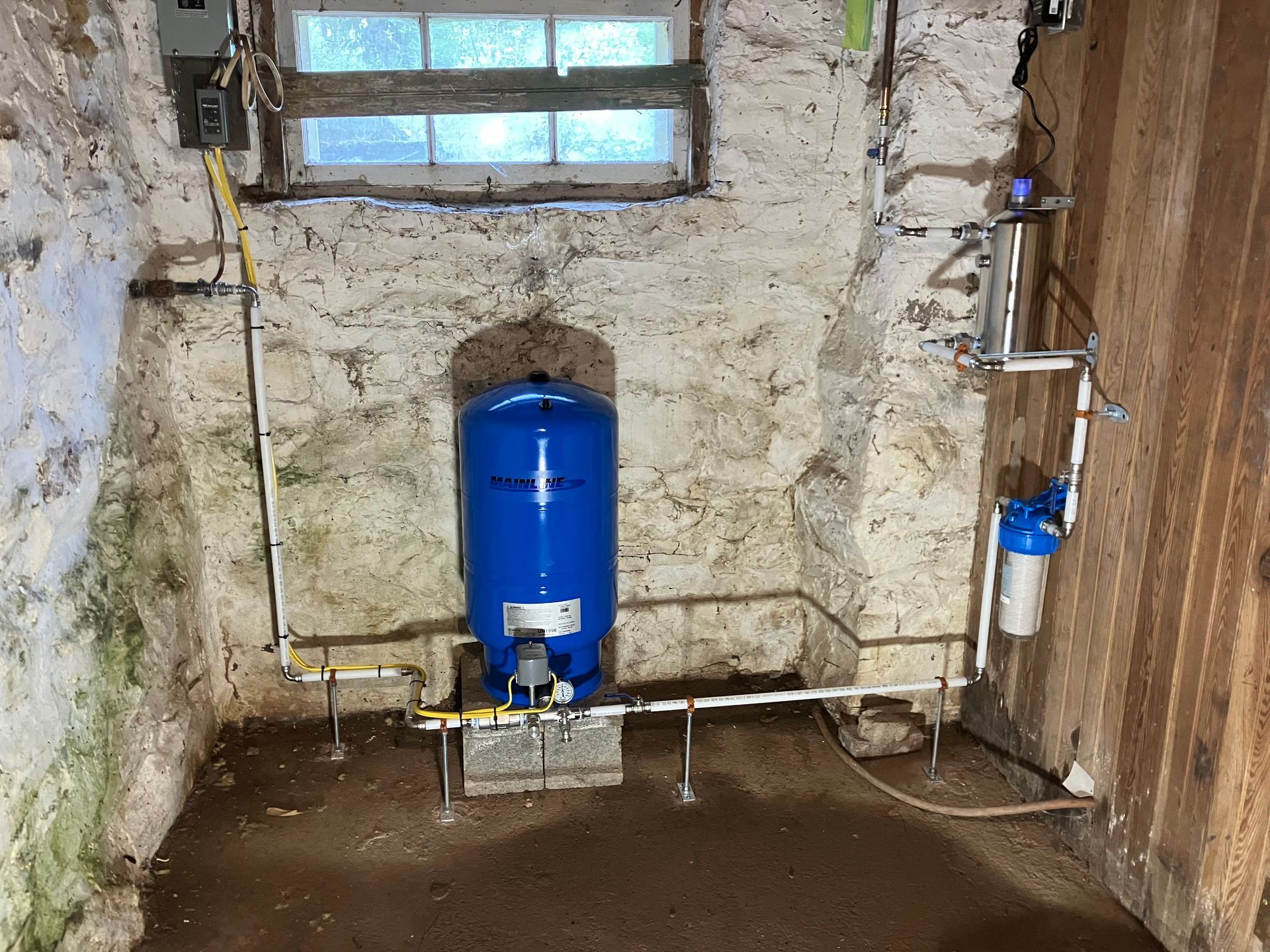 Basement with blue water tank, pipes, window, and filter on stone and wood walls.