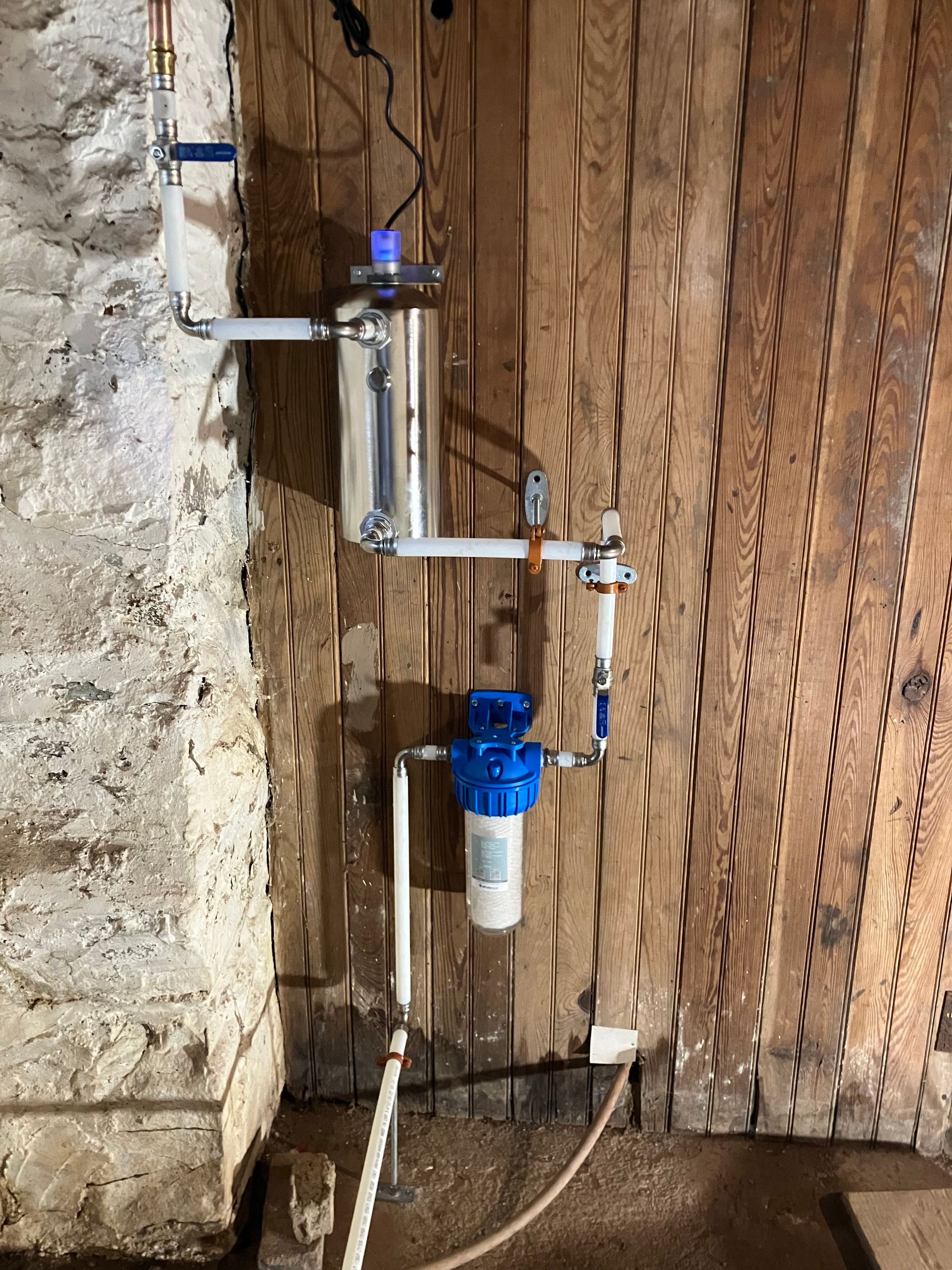 Water filtration system mounted on a wooden wall. A blue filter housing is connected to pipes.