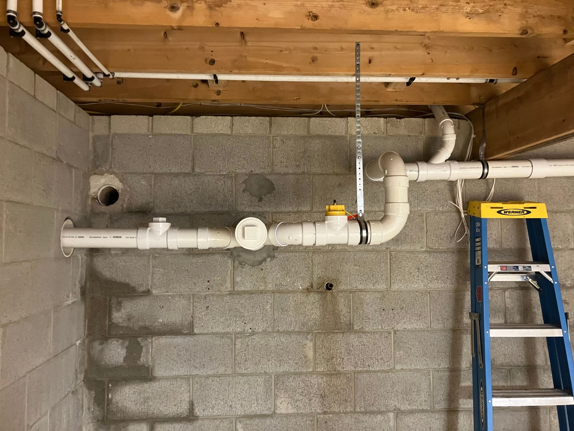 Plumbing pipes and fittings on a gray cinder block wall. A ladder is on the right.