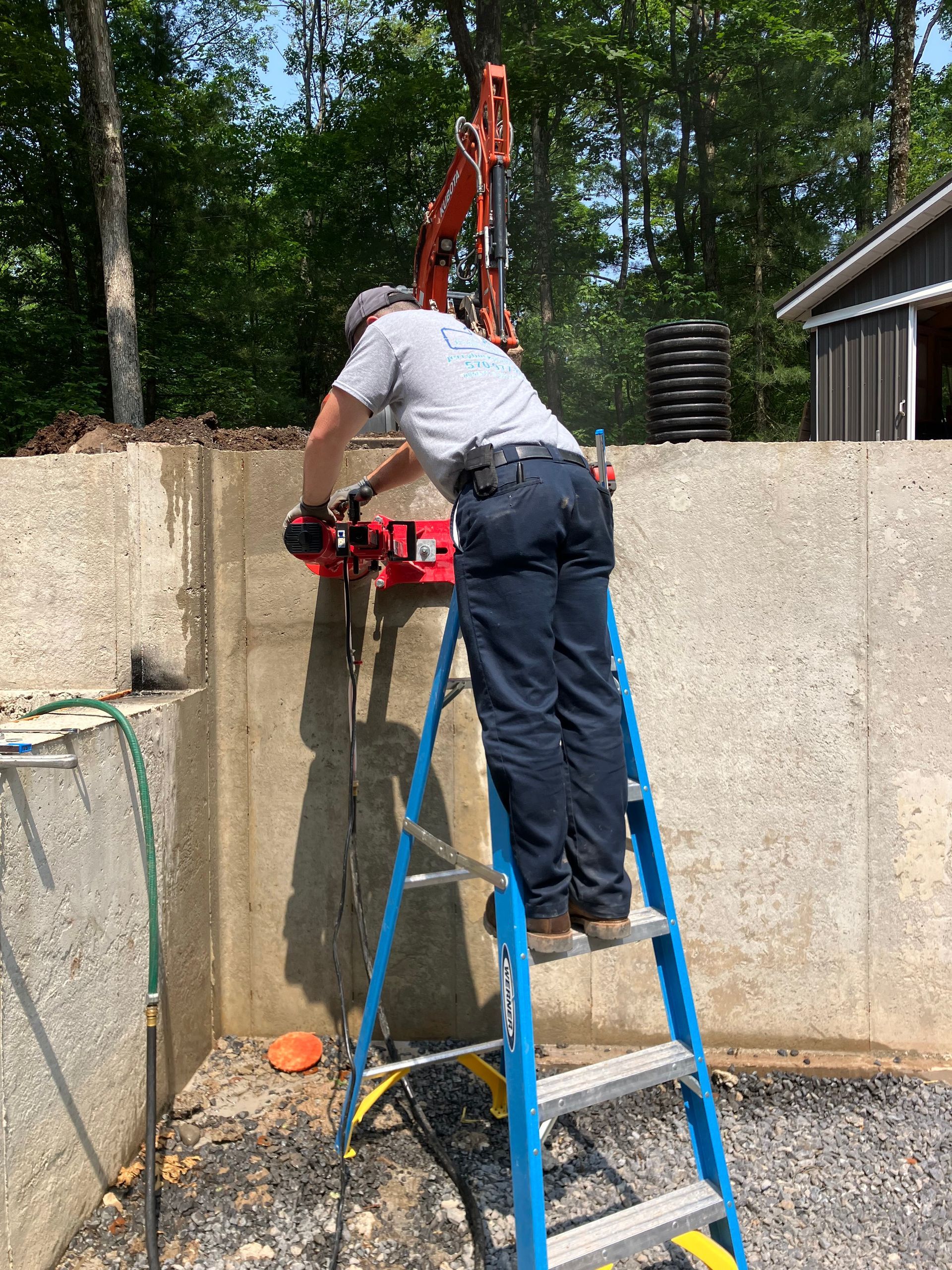 Man on ladder cutting concrete wall with saw outdoors.