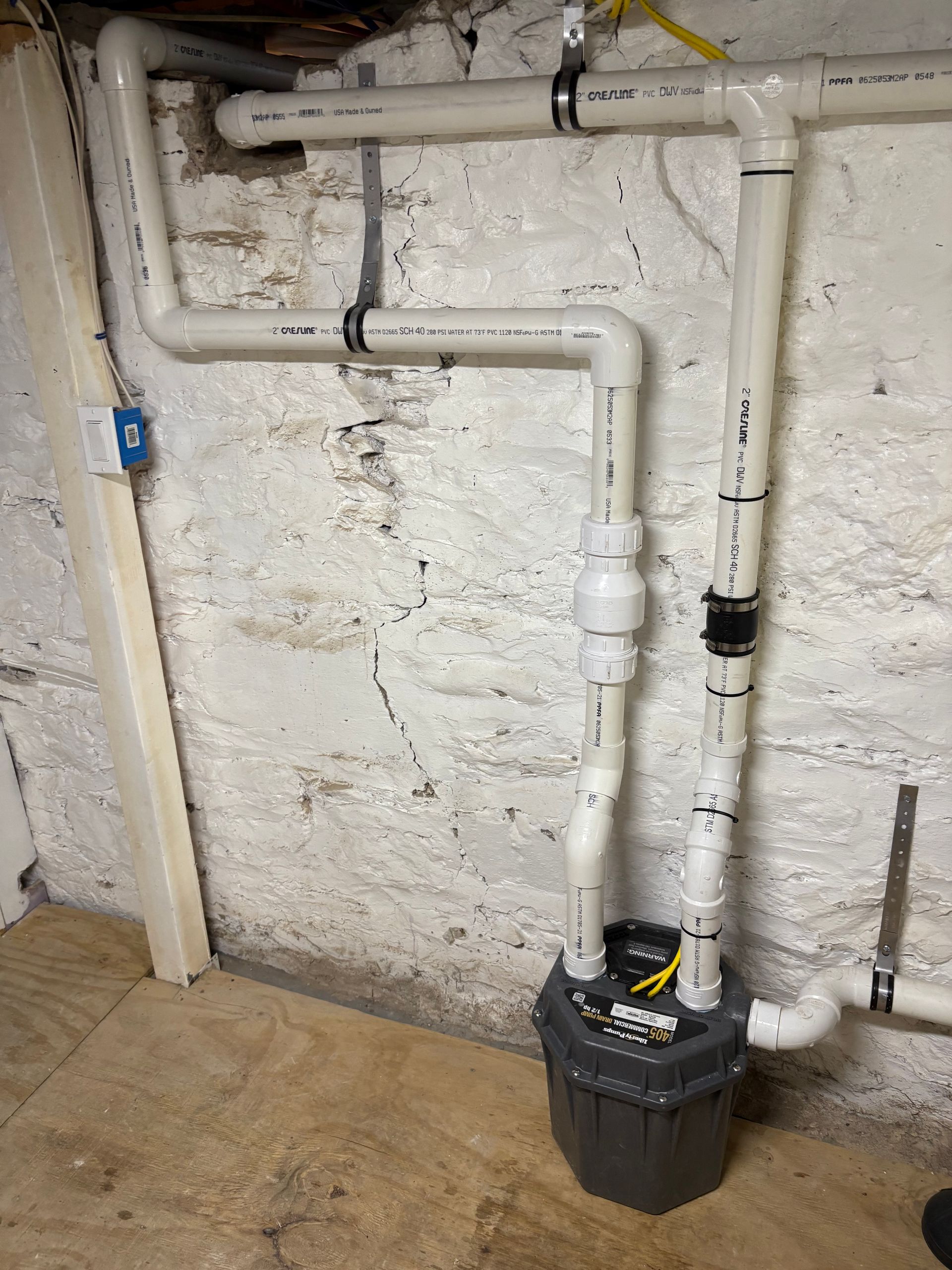 Sump pump system with white PVC pipes against a textured white wall. Gray pump container sits on the floor.