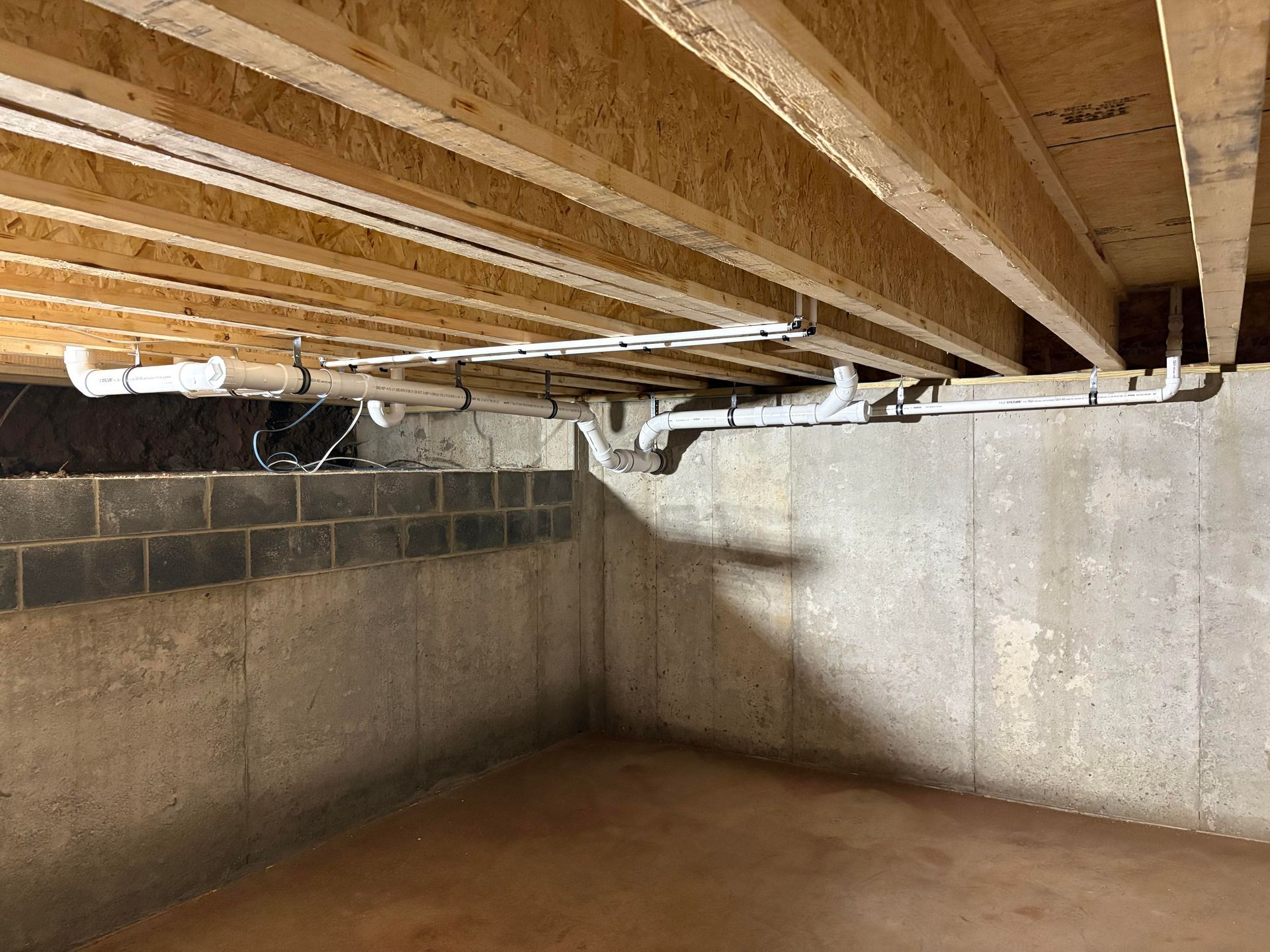 Basement with exposed wood beams, plumbing pipes, concrete block wall, and floor.