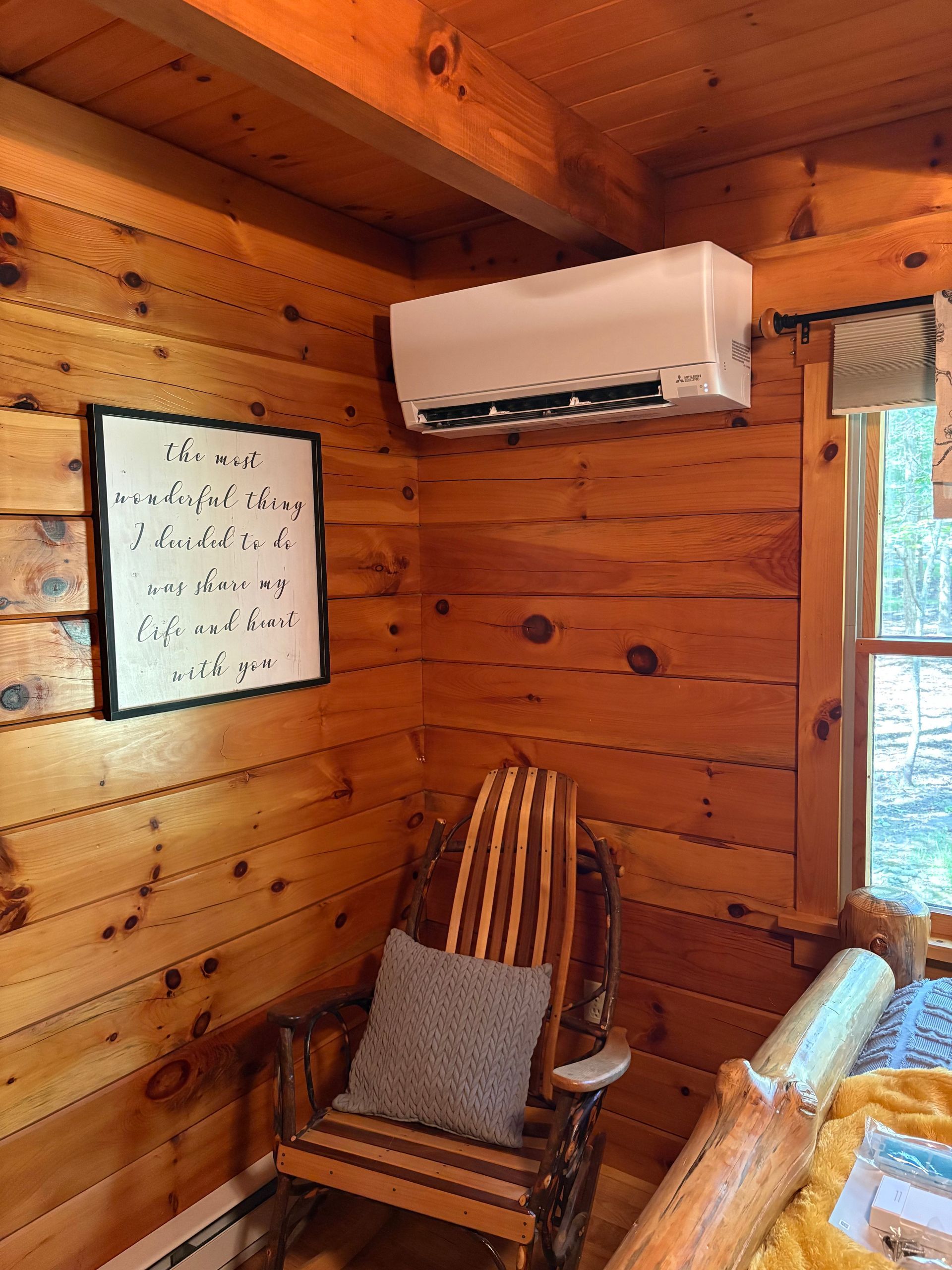 Cozy cabin interior with air conditioner, framed quote, wooden rocking chair, and window. Wooden walls.