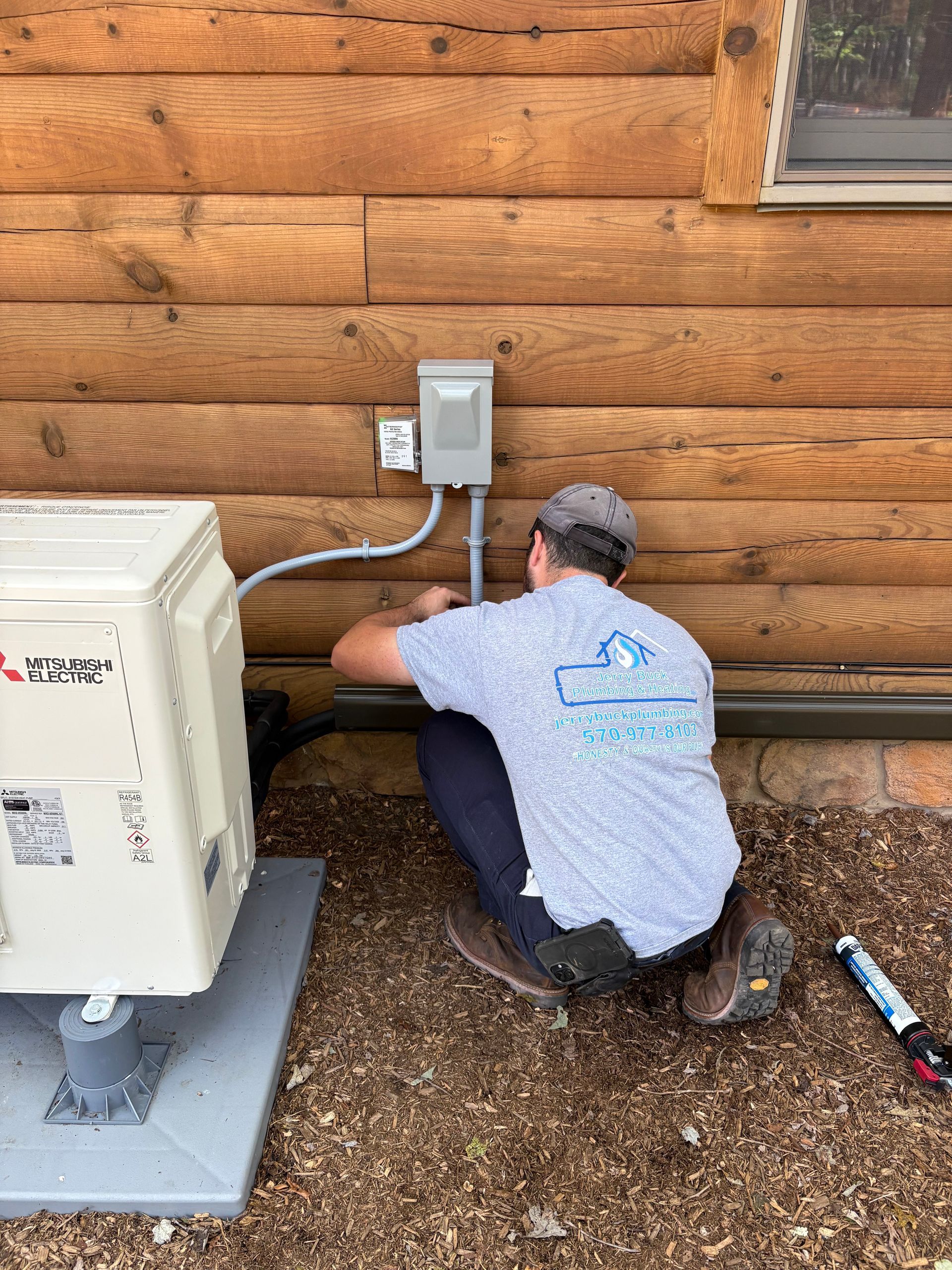 An electrician working on wiring outdoors by a log cabin wall and HVAC unit.