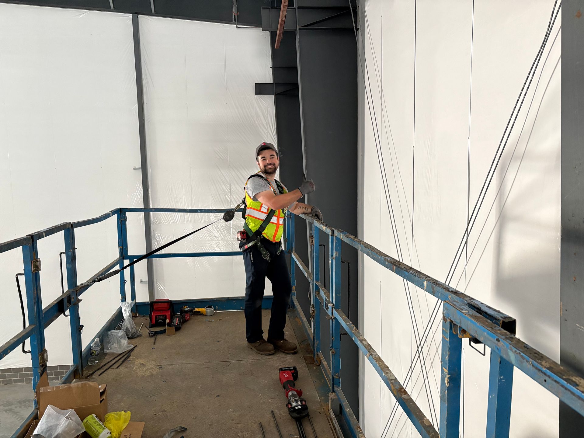 Man on a lift platform, wearing a harness, working on a wall inside a building.
