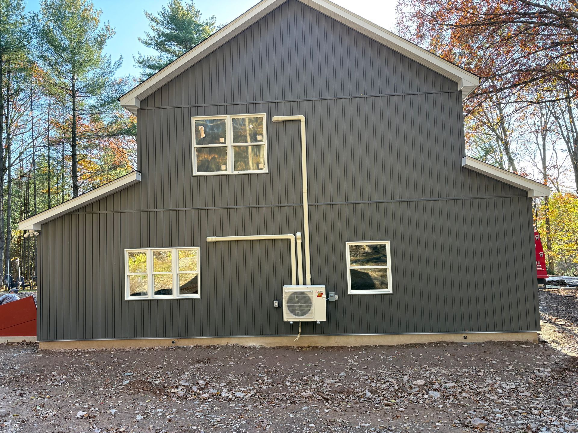 Gray-sided house with three windows, outdoor HVAC unit, and white pipes on a dirt lot.