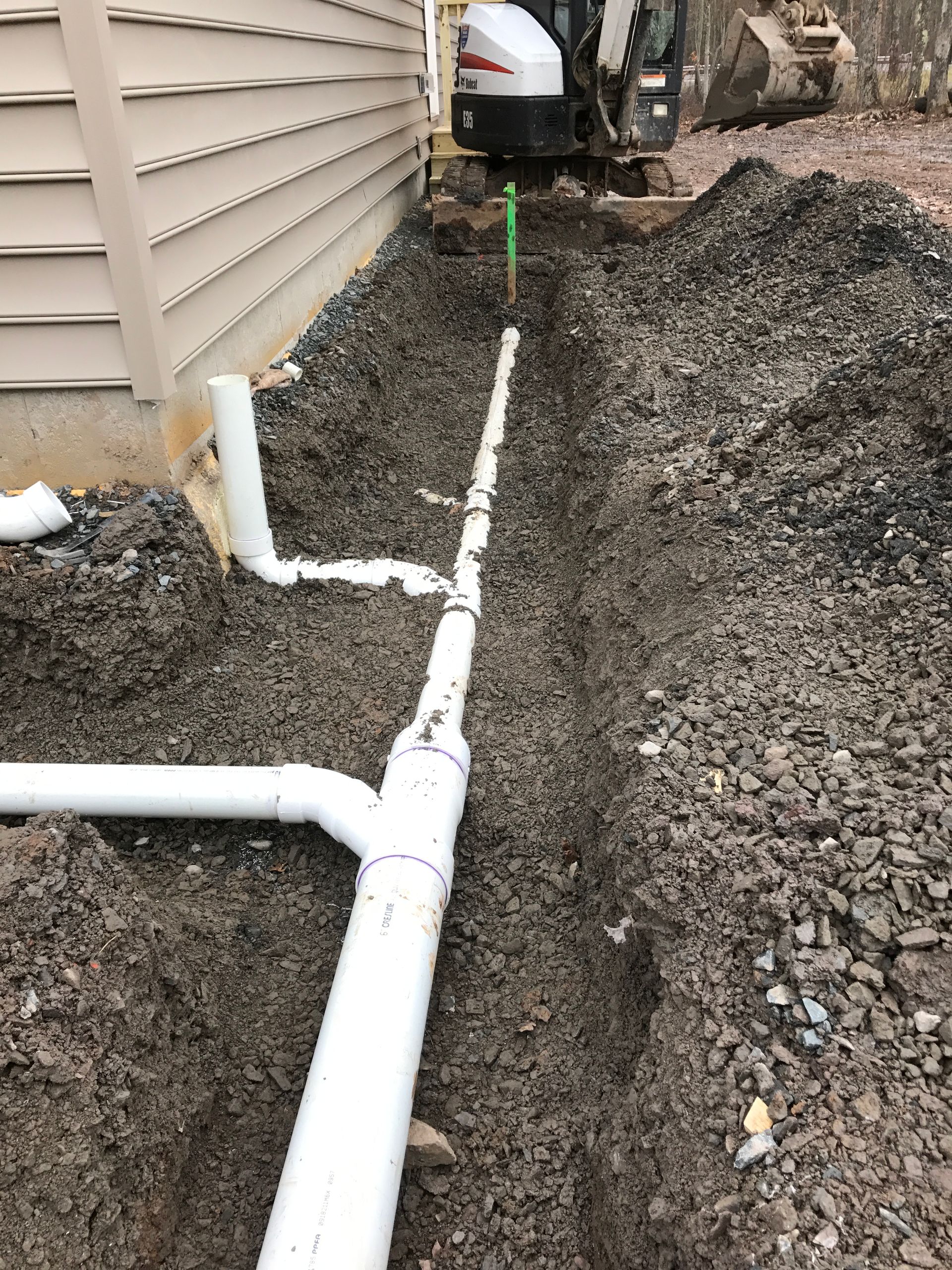 White PVC pipes in a trench near a house foundation, with a backhoe in the background.