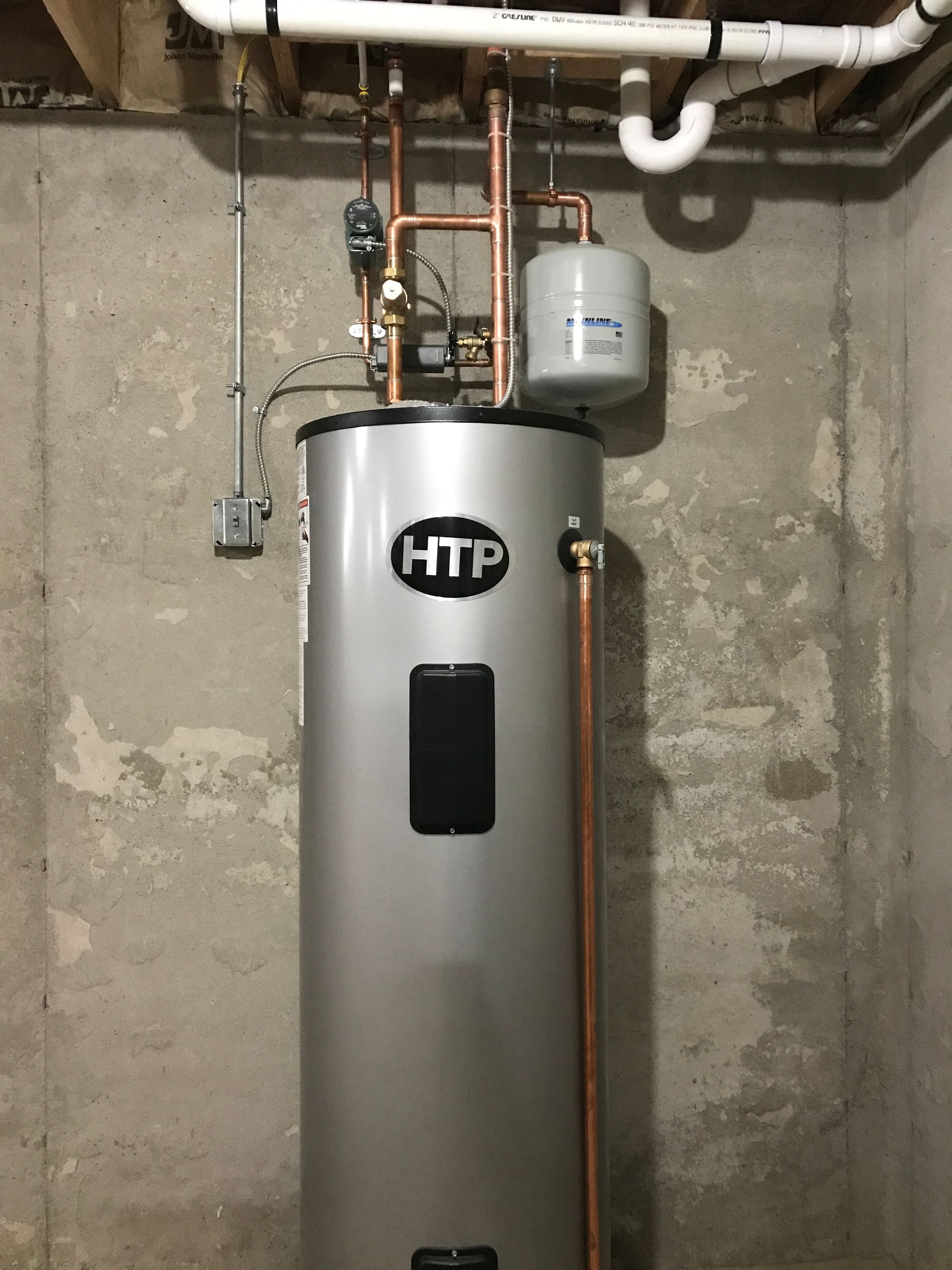 Silver HTP water heater in a basement, with copper pipes, a gray expansion tank, and concrete walls.