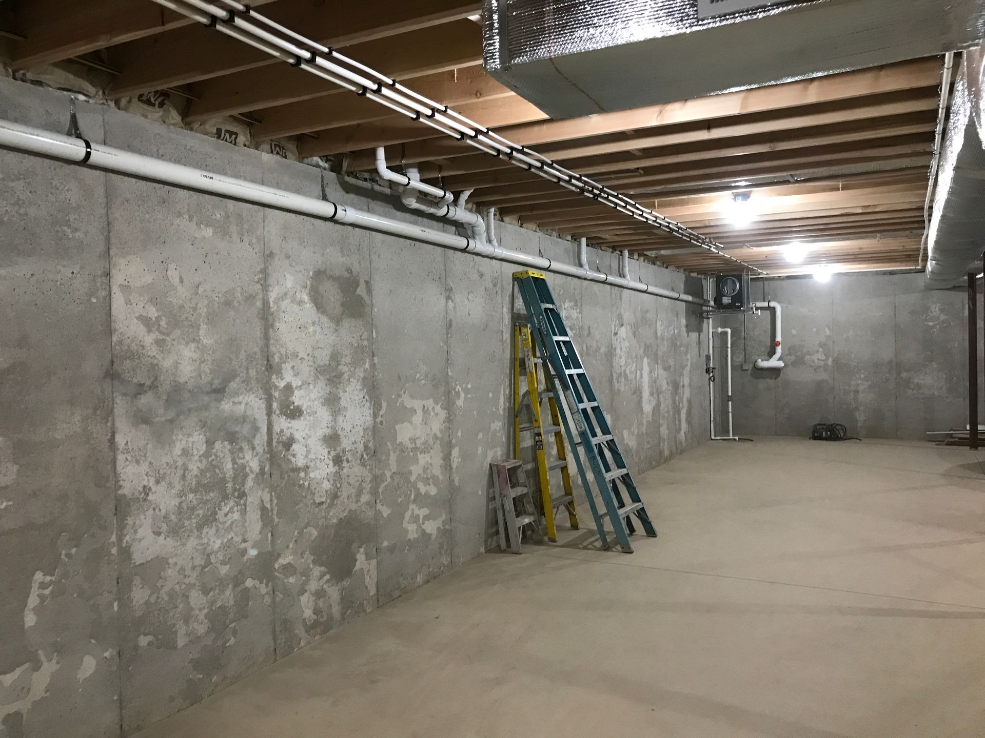 Unfinished basement with concrete walls, pipes, and ladder. Ceiling has exposed wood beams and ductwork.