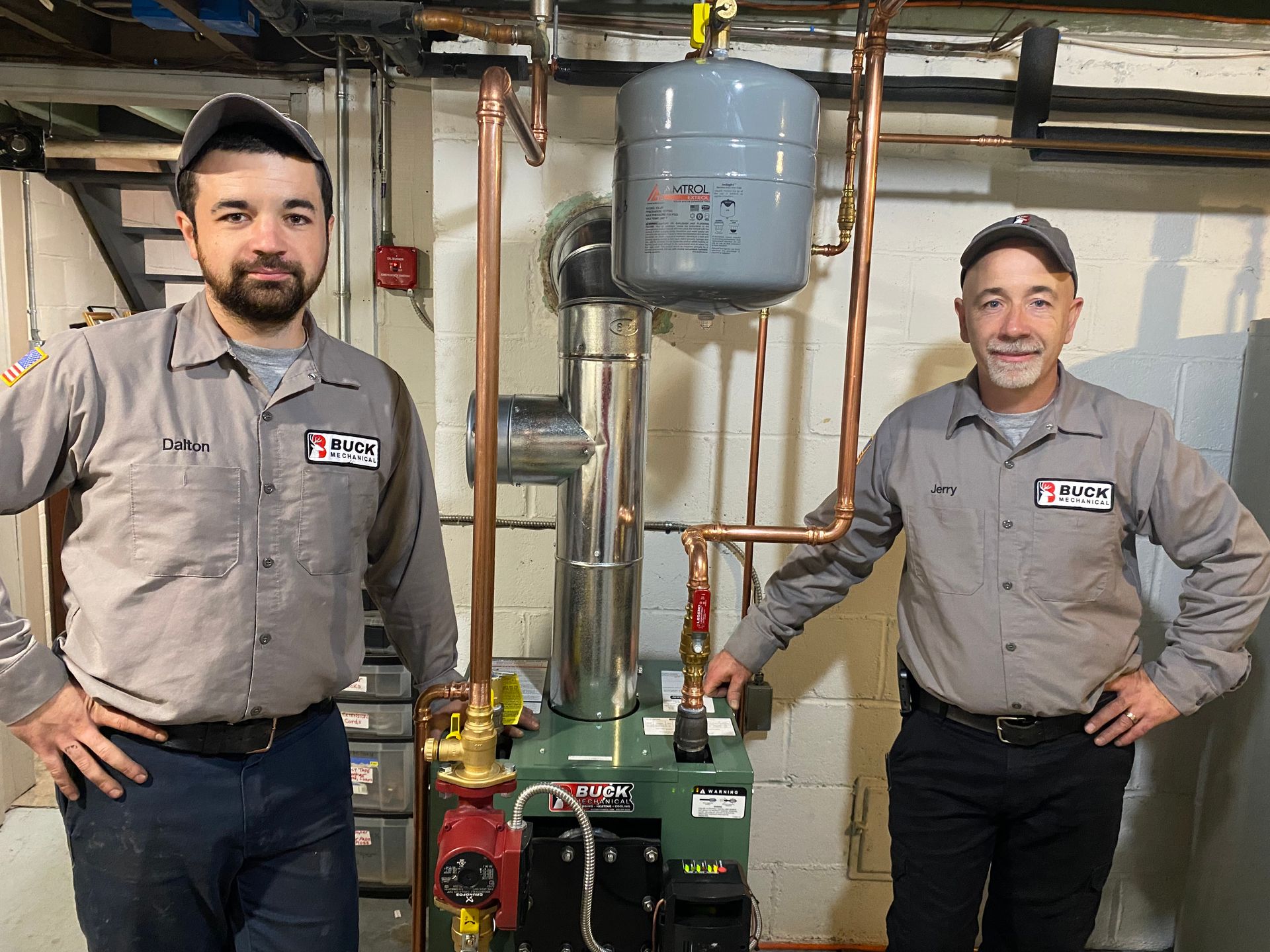 Two technicians pose in front of newly installed plumbing equipment in a basement.