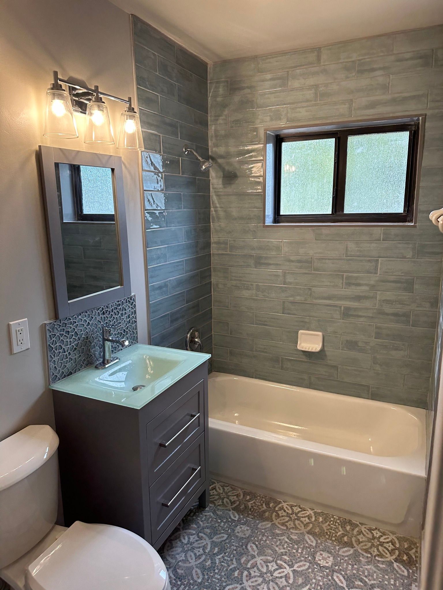 Small bathroom with blue-green tiled walls, gray vanity, patterned floor tiles, and a window over the bathtub.