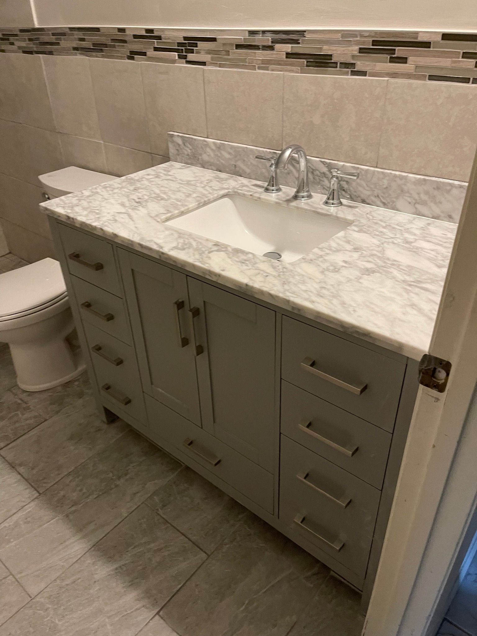 Gray bathroom vanity with marble countertop, sink, and drawers; toilet on the left.