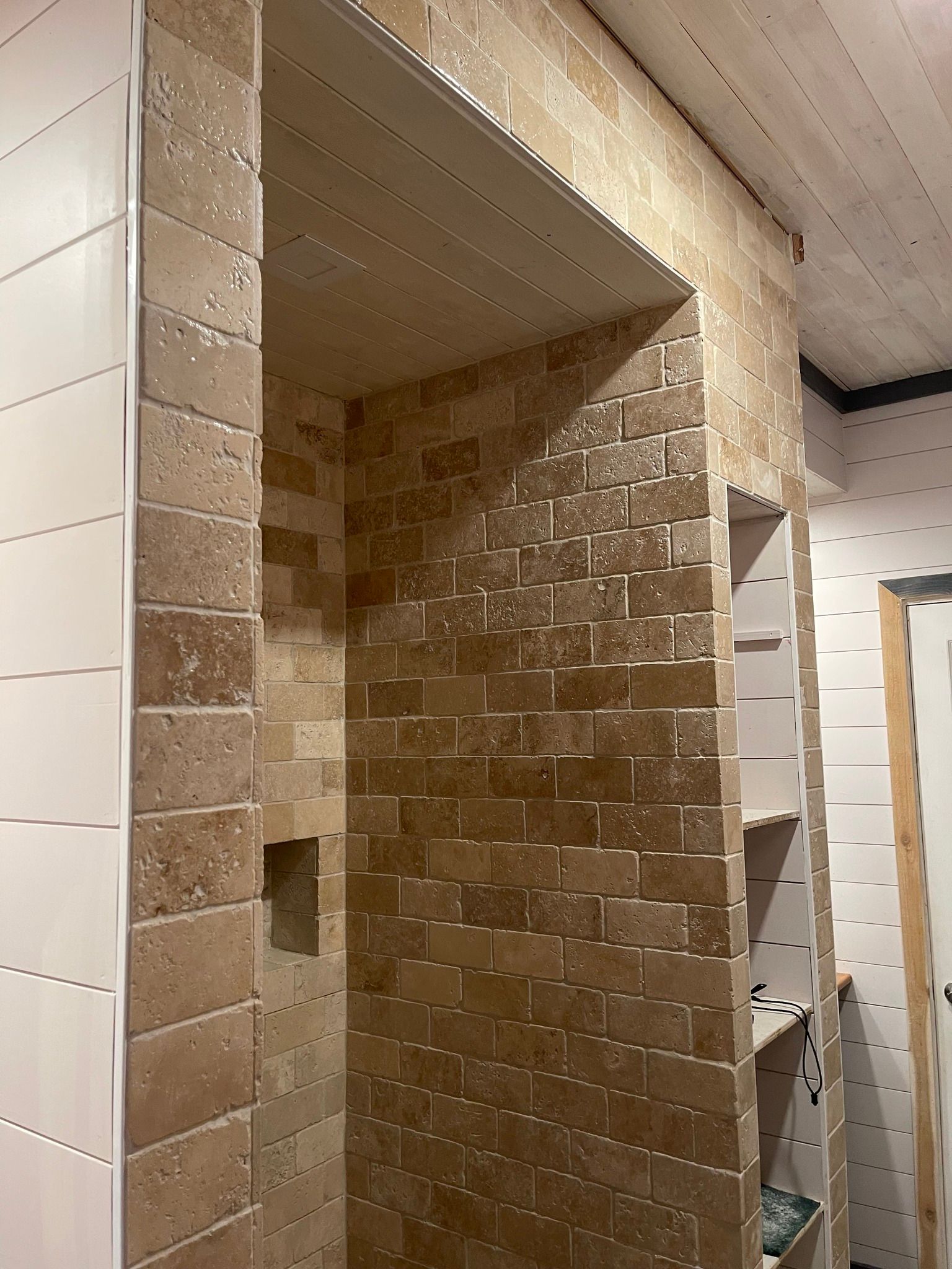 Shower with tan brick-like tile walls and white wood paneling, with built-in shelves on the side.