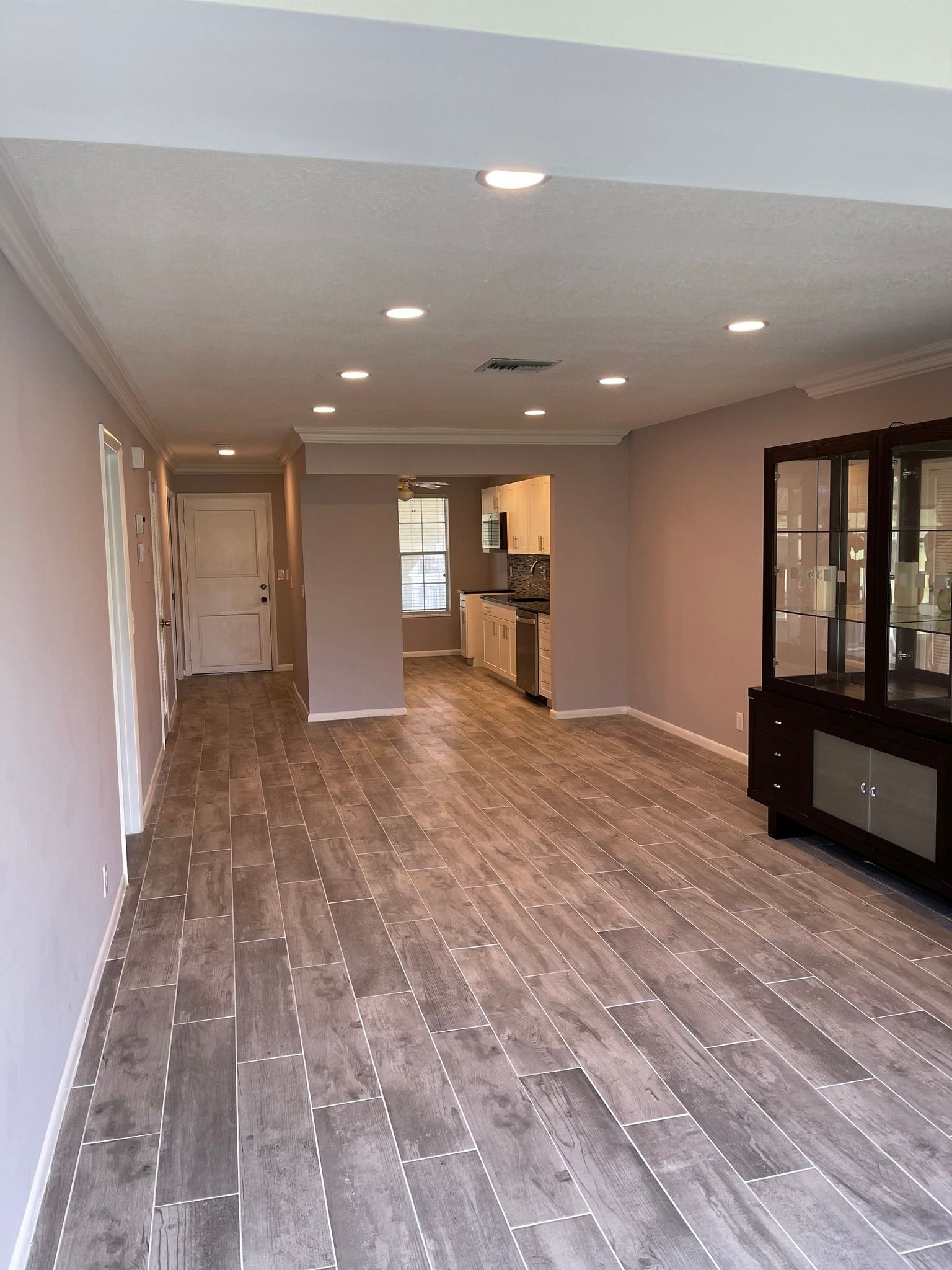 An empty, open living space with wood-look flooring, lilac walls, and recessed ceiling lights; a dark cabinet is to the right.