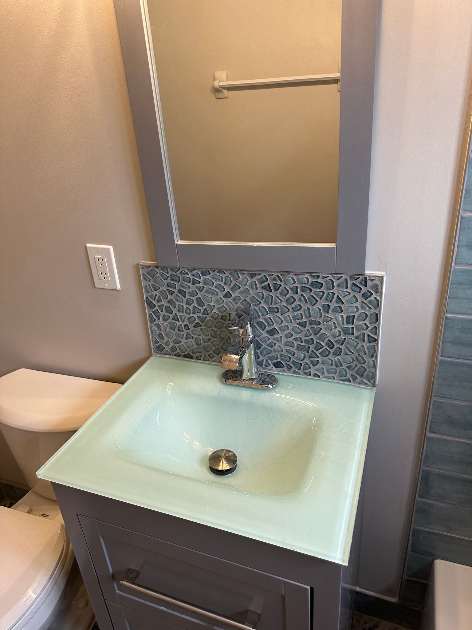 Gray bathroom vanity with a glass sink, mirror, and mosaic tile backsplash.