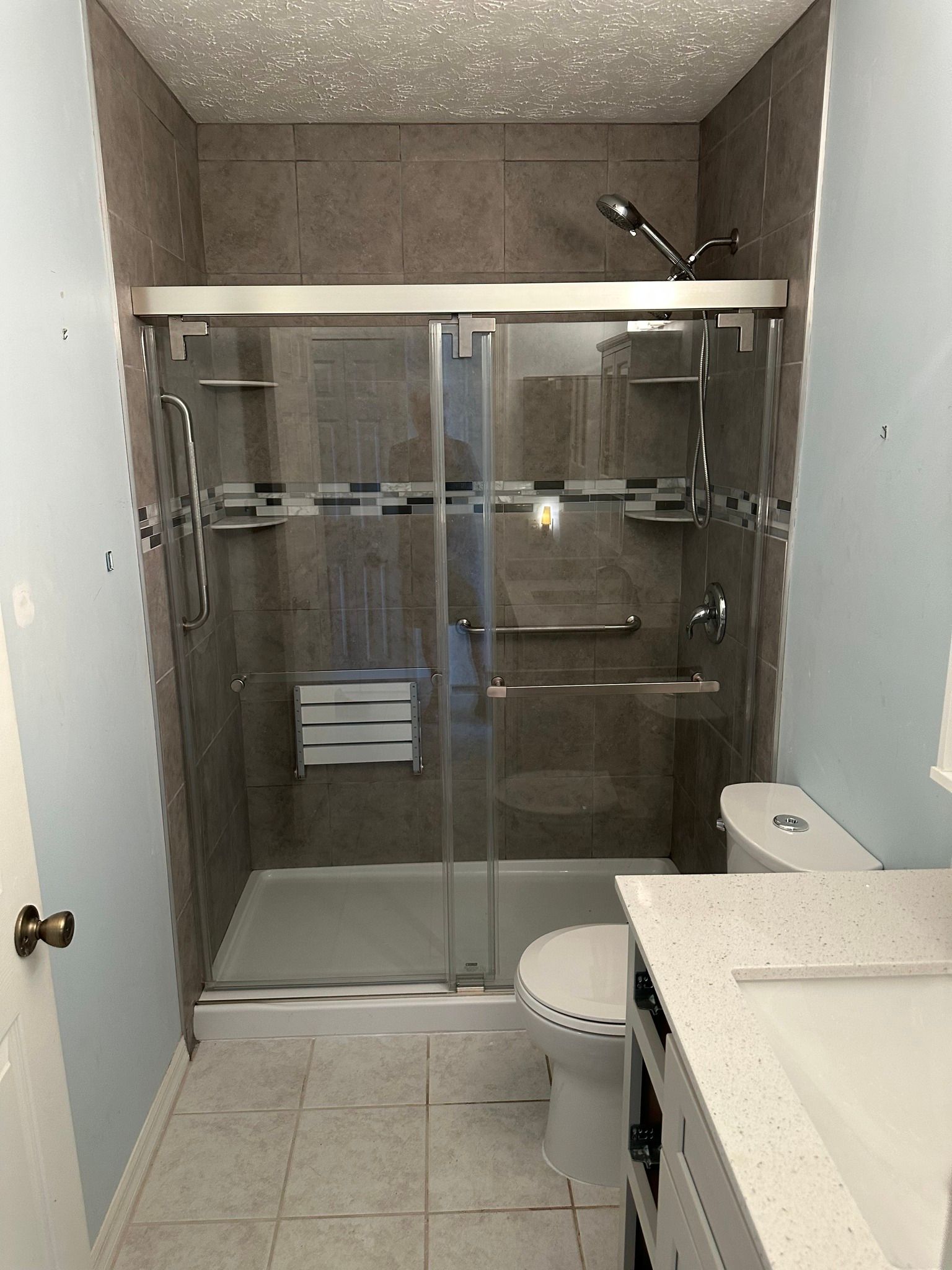 Bathroom with gray tiled shower and glass doors, toilet, and light blue walls.