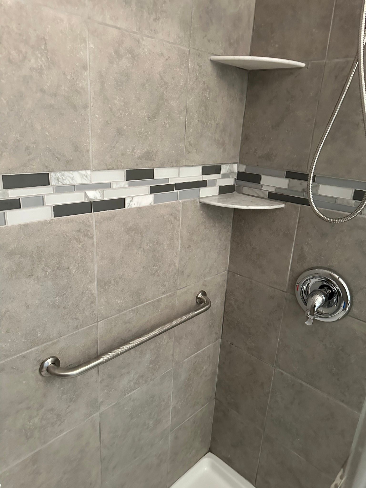 Shower with gray tiled walls, decorative mosaic stripe, shelves, towel bar, and chrome fixtures.