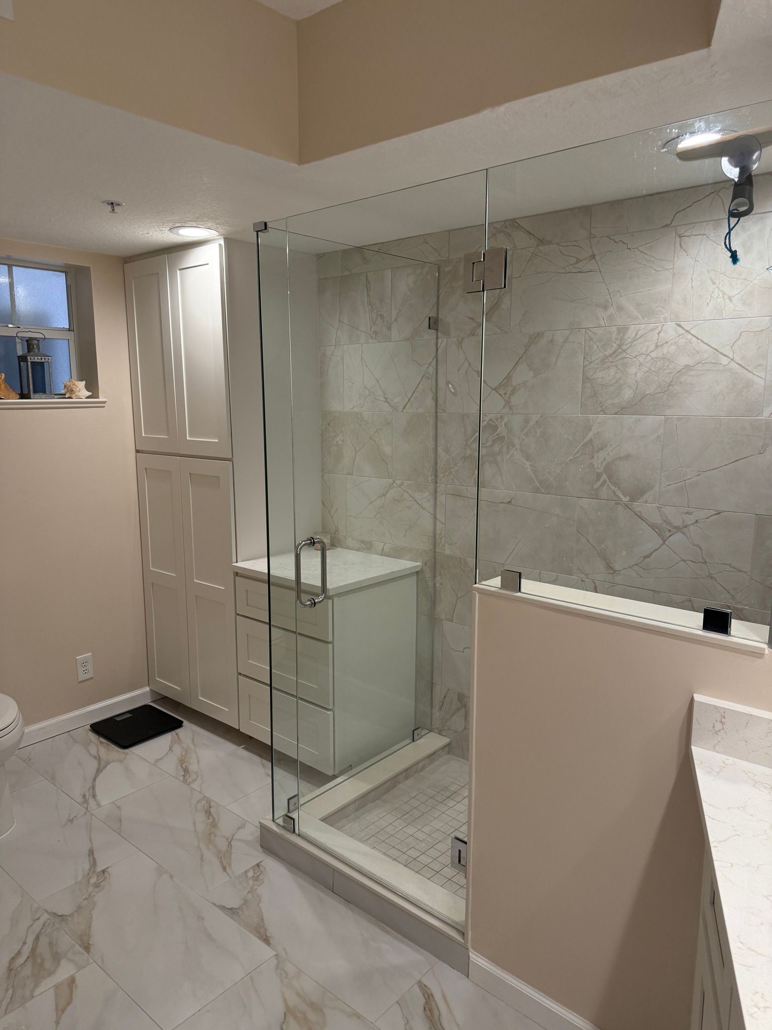 Modern bathroom with marble floors, glass shower, white cabinets, and tan walls.