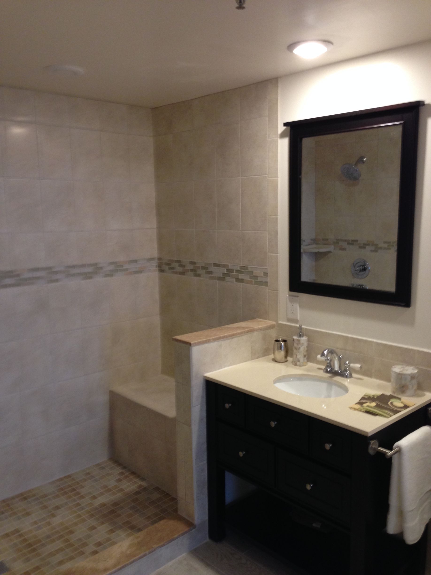 Bathroom with tan tiles, black vanity, mirror, and shower with a bench.