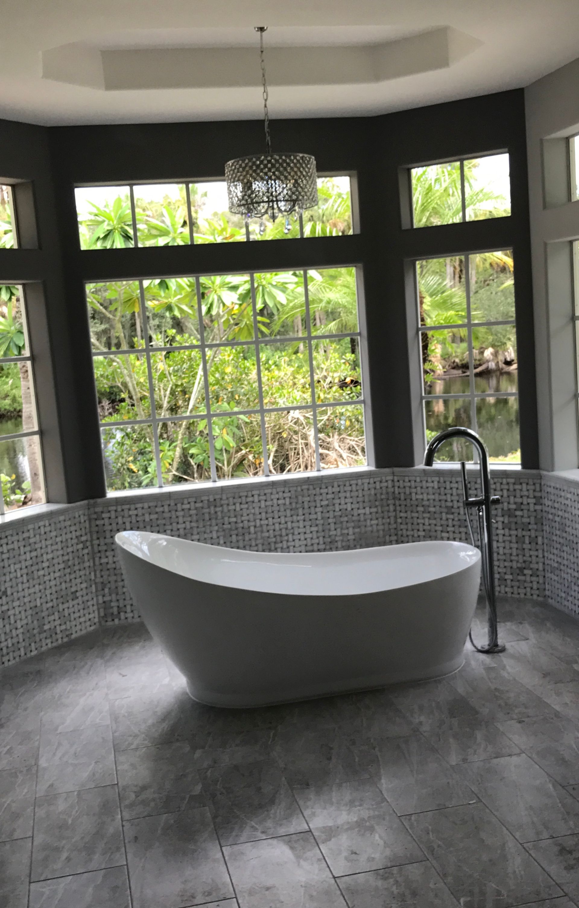 Modern white bathtub in a bathroom with large windows and a chandelier; grey and white tile.